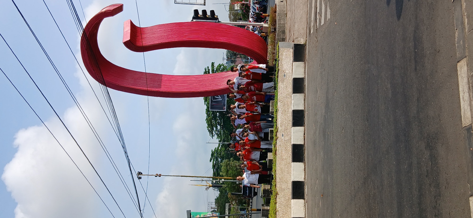 A group of people, some wearing red and white shirts, stand in front of a large red sculpture. The sculpture is abstract and appears to be made of curved, layered materials. Behind the sculpture, a traffic light is visible. The scene is set outdoors, with a bright sky and some clouds. The sculpture is located in the city of Samarinda, Indonesia, as indicated by the context provided. The people appear to be posing for a photograph. The ground in front of them is paved, and there is a low, decorative wall with black and white stripes. Trees and buildings are visible in the background, along with a street and parked vehicles. The overall impression is of a public space with a prominent art installation.