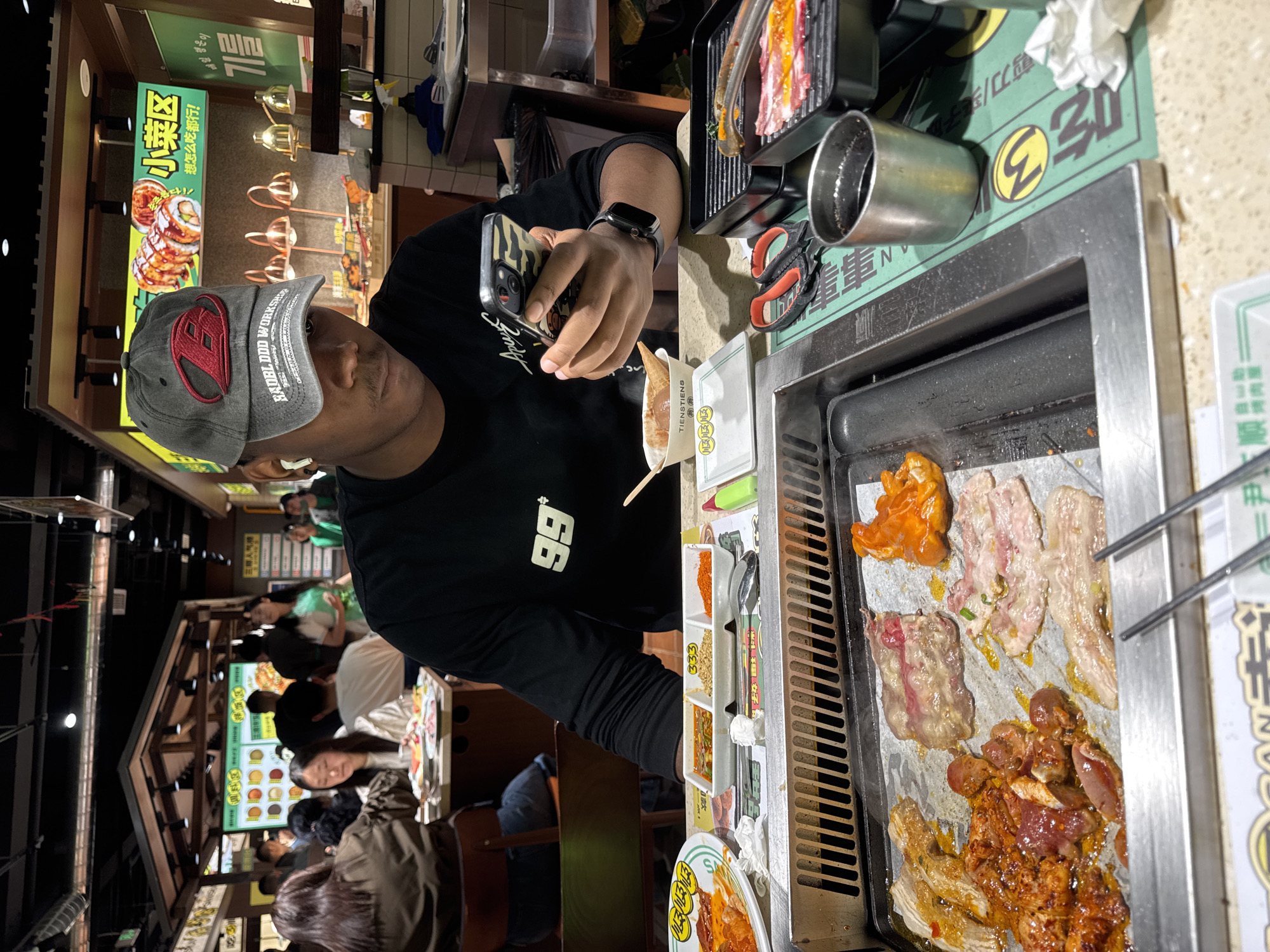 Here is a description of the image:
A person is seated at a table in a busy restaurant, likely in the Chenghua District of China. The individual is taking a photo with a smartphone, possibly of the food being cooked on the grill in front of them. The grill is loaded with various types of meats and food items. Nearby are condiments and utensils, including scissors. A small ice cream cone sits beside the grill. Other patrons are visible in the background. Signs with Chinese characters are on display, adding to the cultural setting. The person is wearing a black shirt with the number 99 and a cap with "BADBL DDD WORKSHJE" printed on the front.