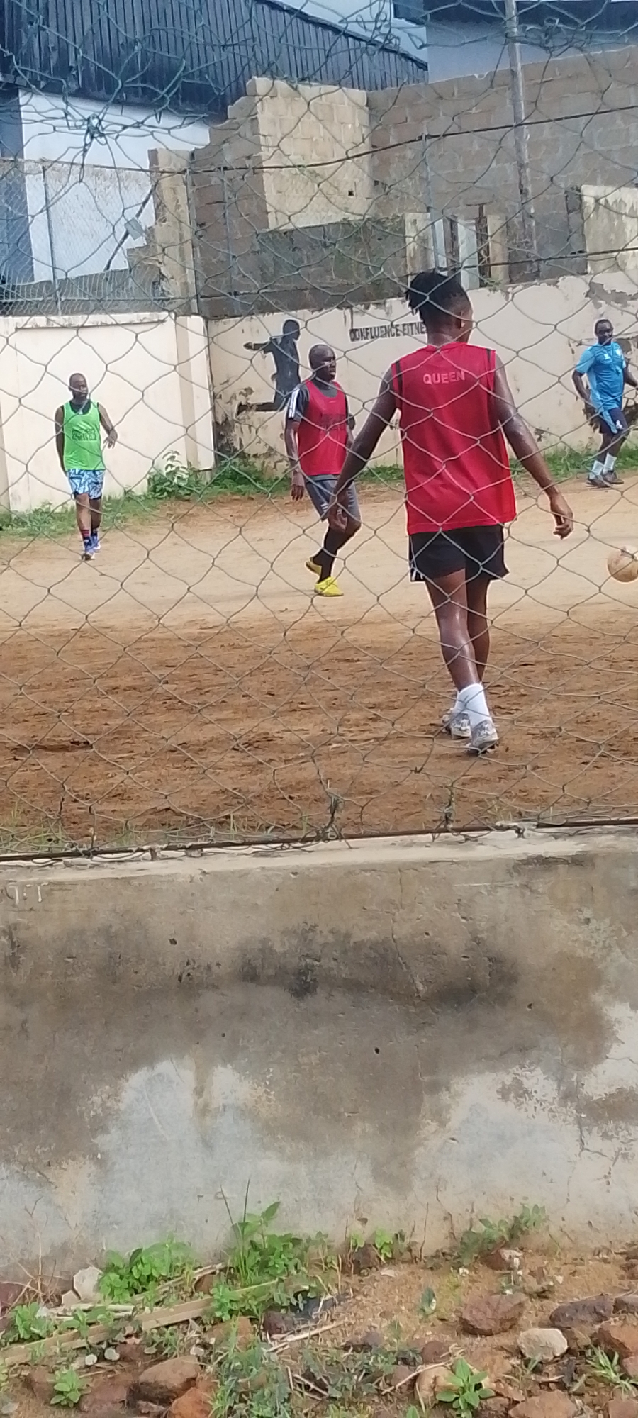 The photo depicts a sports activity likely taking place in Lokoja, Nigeria. The foreground shows a chain-link fence with a concrete base. Behind the fence, several men are engaged in a game on a dirt field. One man, facing away from the camera, wears a red jersey with "QUEEN" printed on the back. Another man wears a green vest and colorful shorts. Others wear blue and red vests. In the background, there is a building with partial ruins or damage visible. The building's wall has writing on it, including the words "CONFLUENCE FITNESS." The overall scene appears to be a casual game in an outdoor setting.