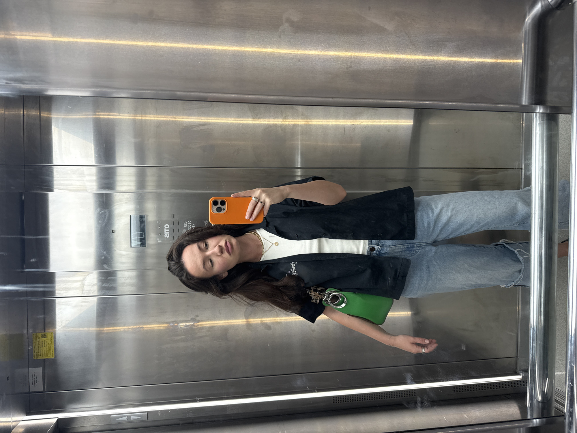 The image shows a young woman taking a selfie in an elevator with stainless steel walls. She is wearing a white top, a black shirt, light blue jeans with a ripped knee, and a gold chain necklace. She is holding an orange mobile phone in her left hand. A small green purse is attached to her shirt. The elevator has a control panel with buttons and signage in Hebrew. The image was taken in Tel-Aviv, Israel.