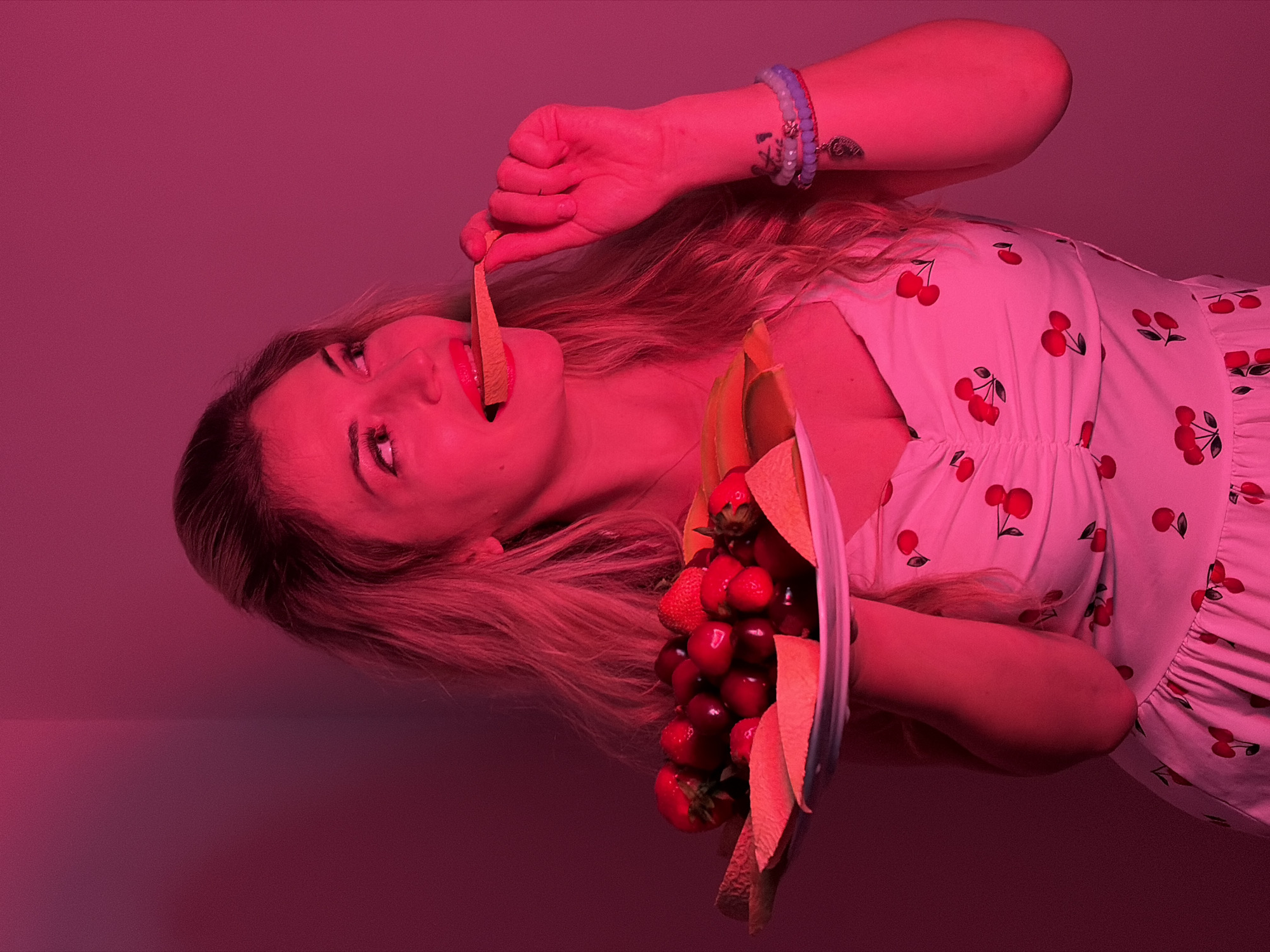 A blonde woman in a cherry-printed sundress is depicted in a pink-toned image. She is holding a bouquet of strawberries and cherries, and puts an orange slice in her mouth. The woman has a bracelet on her left wrist. The scene lacks distinct spatial context, therefore a location in Budva, Montenegro cannot be determined.