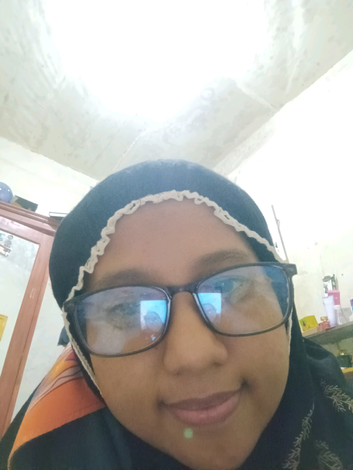 A close-up, front-facing selfie of a young woman wearing a black hijab and glasses is captured. She is looking towards the camera with a slight smile. The background is blurry, but suggests an indoor setting, possibly a room with furniture and items on shelves, including bottles and boxes. A wardrobe or cabinet with a glass door is visible on the left side of the frame. The lighting is bright, with the ceiling appearing to be the primary light source. The image does not contain any specific geographical markers to confirm its location within Sarangan, Indonesia.
