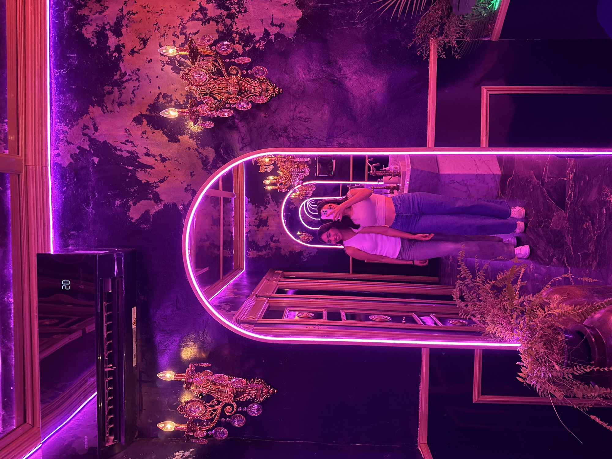 A room with purple neon lighting is shown. There are two girls in a mirror who are taking a selfie. The location of this photograph is in Puebla, Mexico. The walls of the room are a dark purple with gold accents. There are ornate chandeliers on the wall. A black air conditioning unit is on the left wall. There are also plants on the right wall.