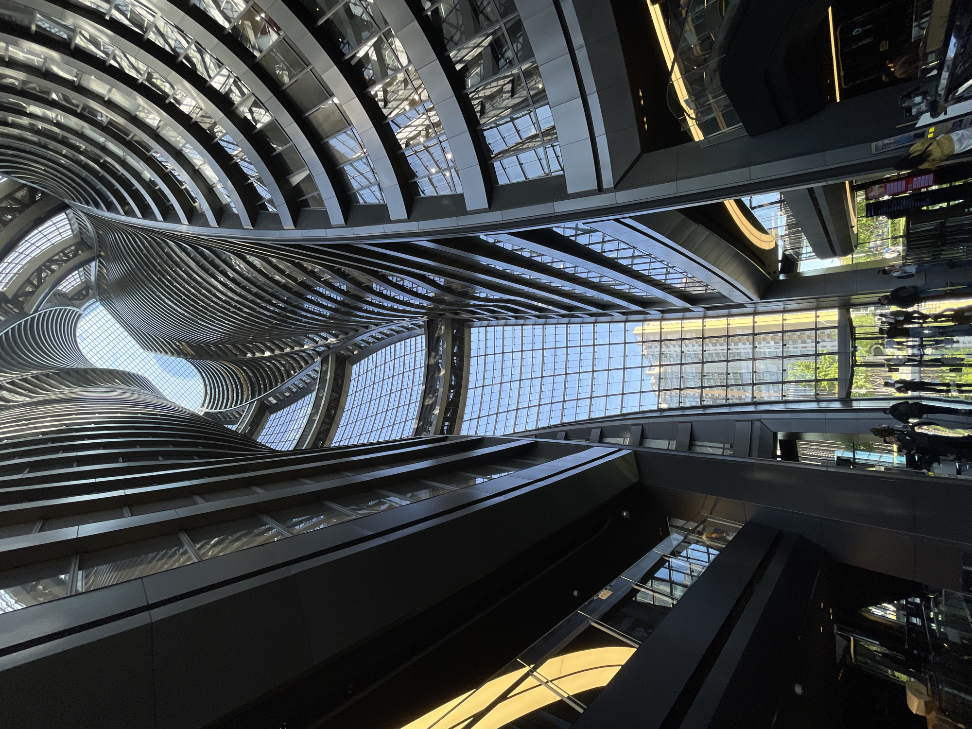 The image depicts an upward-angled view of an architectural structure in Fengtai District, China. The building features an interior with curved, layered tiers and prominent glass panels that allow natural light to filter in. The design incorporates linear elements and reflective surfaces. Reflected figures are visible on the right side of the image, suggesting a public space or walkway. The architectural style is modern and abstract, emphasizing the flow of light and the play of forms.