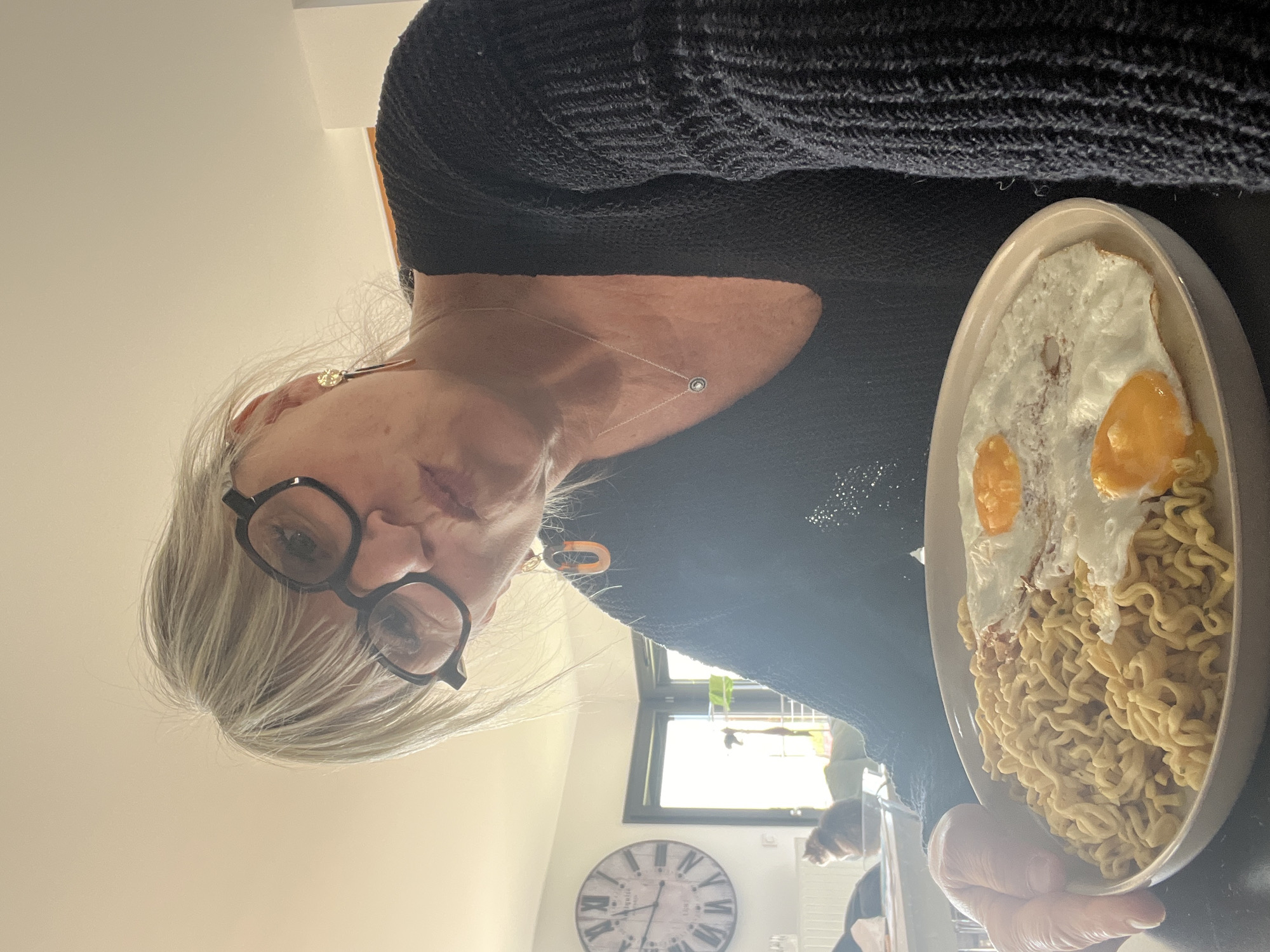 A person with gray hair and glasses is holding a plate of noodles with two fried eggs. The person is wearing a black sweater and a necklace. In the background, there is a window, a clock on the wall, and another person or object is partially visible. The location appears to be inside a house, possibly in Chambœuf, France.