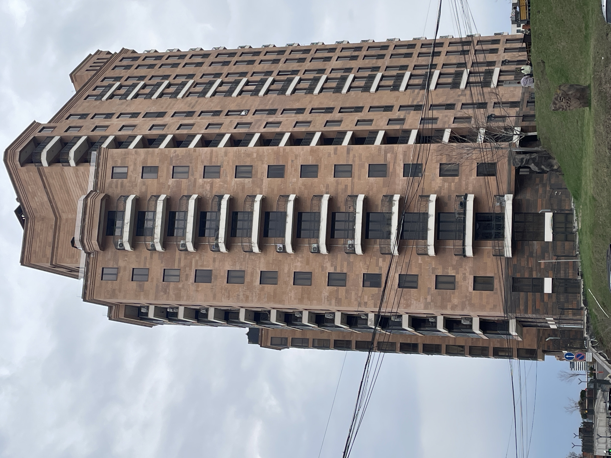 The image shows a tall, brown, rectangular building in Yerevan, Armenia. The building has many stories, each with regularly spaced windows. Some of the stories have balconies with white railings. There are numerous air conditioning units attached to the building's exterior, particularly on the balconies. Power lines are visible in the foreground, obscuring some of the building. A patch of grass and a street are partially visible on the right side of the image. The sky is overcast.