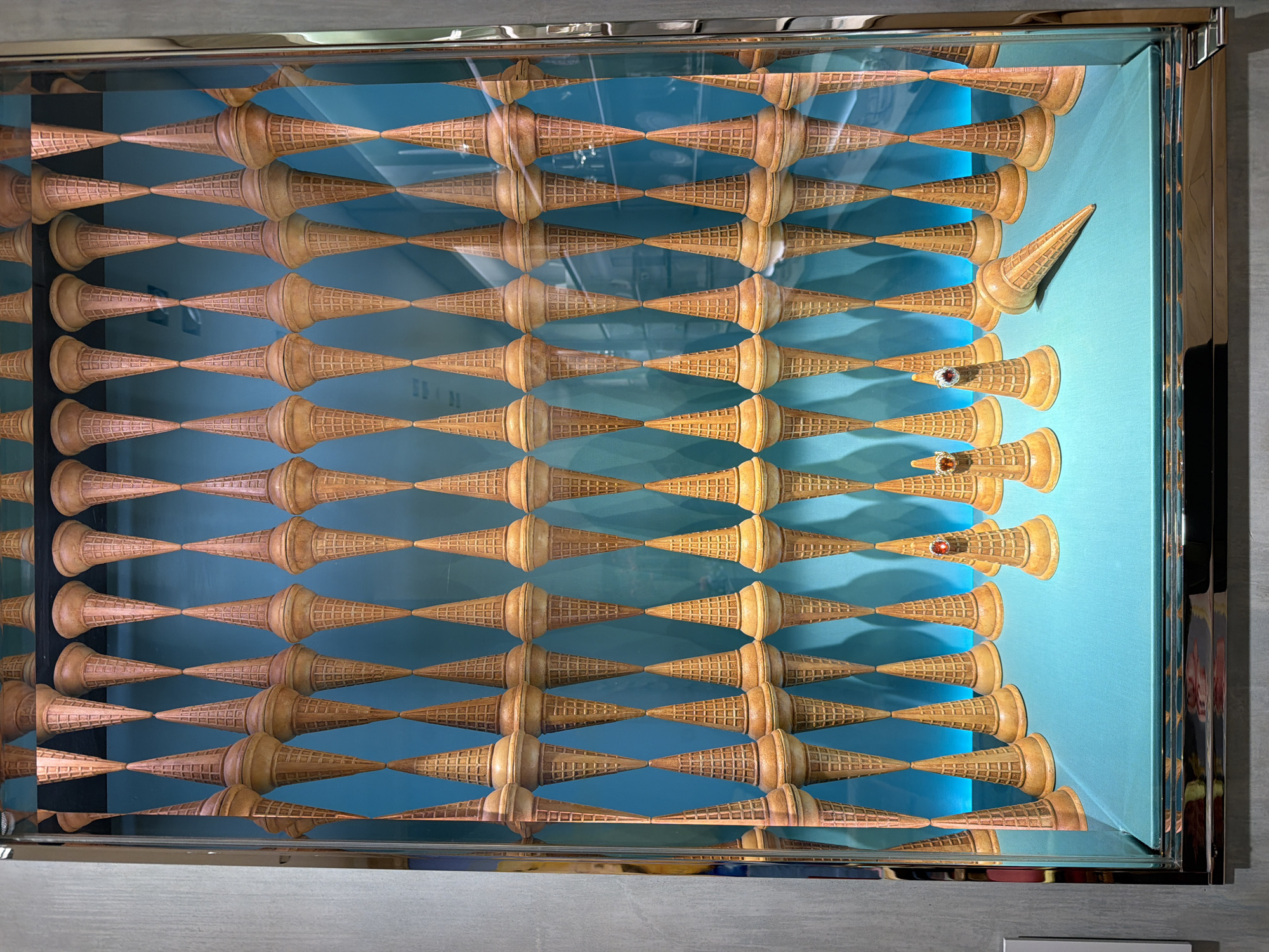 This image is an eye-level, medium shot of framed artwork, depicting an arrangement of ice cream cones. The artwork consists of rows and columns of brown waffle ice cream cones arranged in a grid-like pattern against a turquoise background. Most of the cones are oriented with the open end facing down, while a few on the right side are turned upside down and have rings placed on them. The frame of the artwork is silver and reflective. The background outside the frame appears to be a gray wall. The location is the city of New York, United States of America.