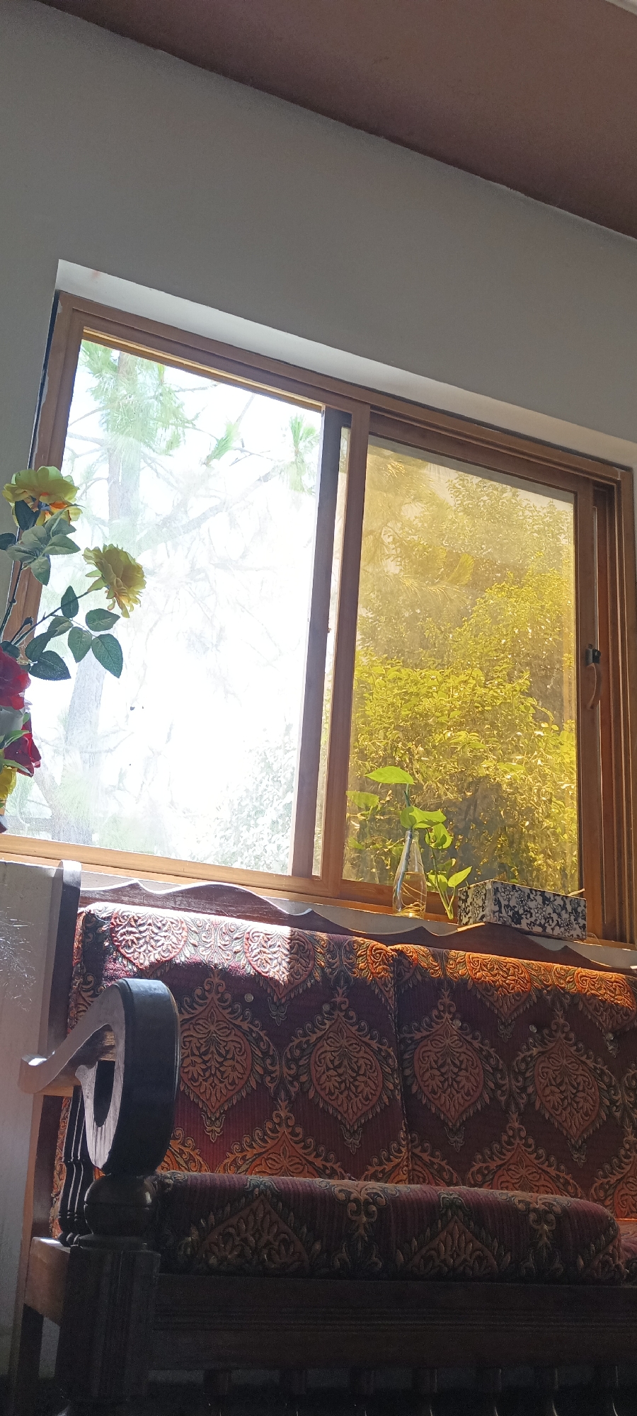 The image depicts an interior scene in a building in Katlang Tehsil, Pakistan. A wooden-framed window with two panes dominates the view. The left pane offers a bright, sunlit view of trees outside, while the right pane presents a view of greenery filtered through a yellowish tint. On the window sill, there are decorative items, including a small vase with green leaves and a patterned container. Below the window, a section of a sofa or couch is visible, covered in a patterned fabric with a burgundy and gold color scheme. A partially visible wooden armrest of the sofa is on the left side of the frame. A bouquet of artificial flowers, predominantly red and yellow, is also positioned near the left side of the window. The ceiling is painted white, with a hint of brown towards the upper edge of the frame.