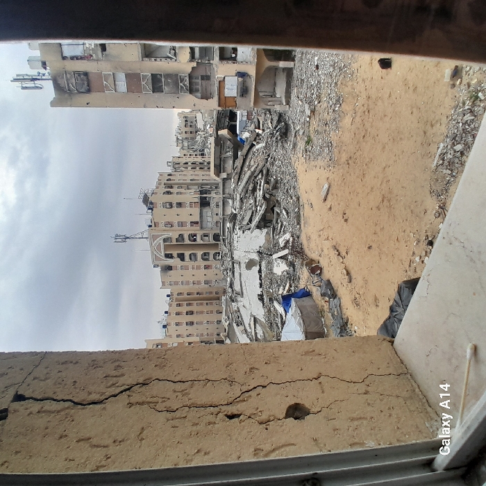 The image depicts a scene of extensive urban destruction in Khan Yunis, Palestinian Territory, viewed from a window. In the immediate foreground, a heavily cracked, light-brown wall or pillar with exposed material is visible on the left, framing the view. A white window sill runs horizontally across the bottom, with a single white ear swab resting on it and a "Galaxy A14" watermark.
Beyond the window, the midground reveals a large expanse of sandy ground littered with significant debris and rubble, primarily concrete fragments and twisted rebar from collapsed structures. Towards the left midground, a rudimentary shelter covered with a blue tarp is present amidst the debris.
The background features numerous multi-story buildings, consistent with residential or commercial architecture, stretching into the distance under an overcast sky. These buildings exhibit widespread damage, including structural compromises, missing sections of walls, exposed interiors, shattered windows, and visible charring and impact marks on their facades. Several rooftops are equipped with telecommunication antennas. No human figures are discernible in the frame.