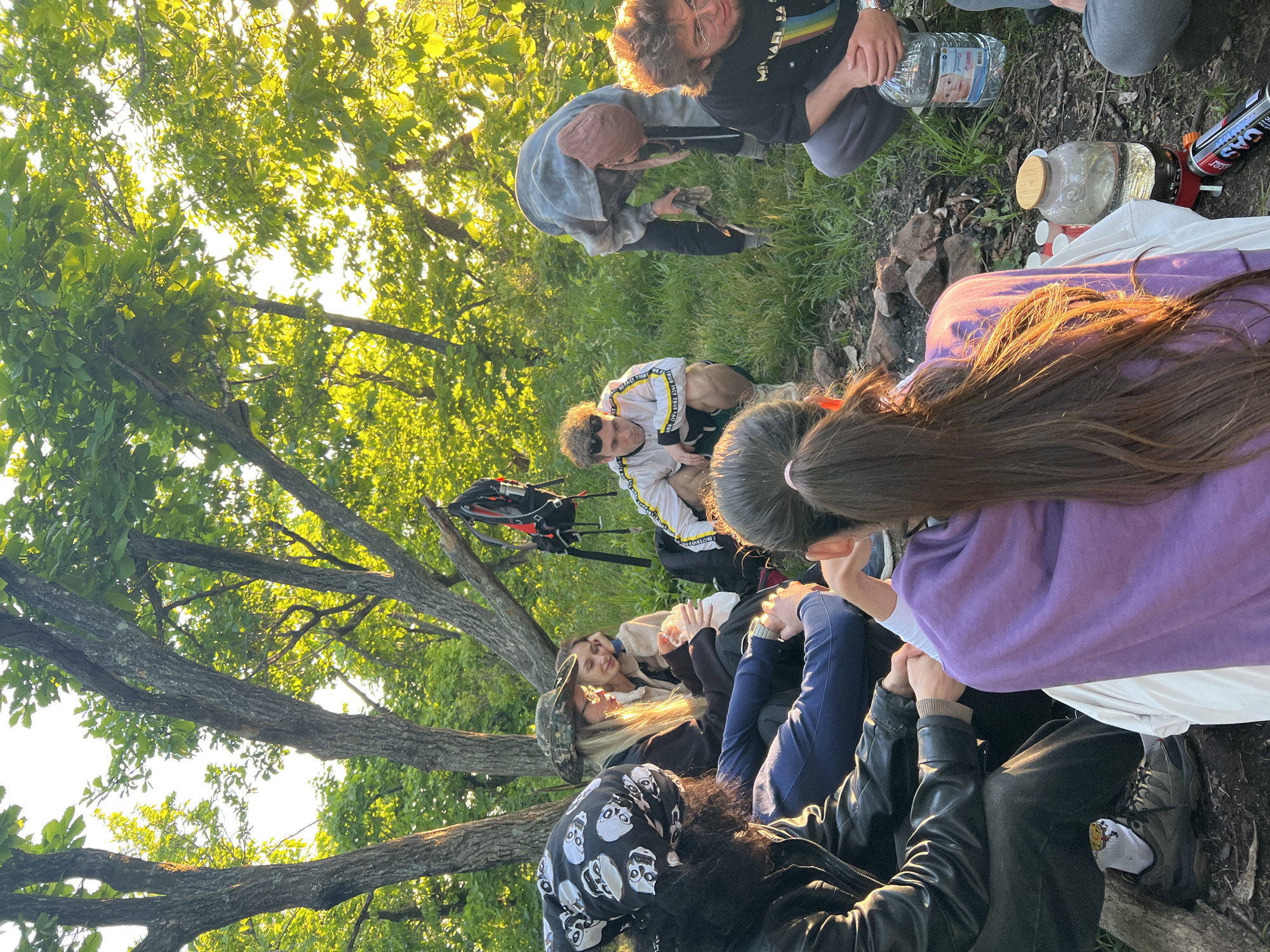 A group of people, approximately 8-10 individuals, are gathered in a circle in a wooded area, possibly a park or forest. They are situated around a makeshift fire pit constructed from stones. Some individuals are sitting on the ground, others are kneeling, and a few are standing. A backpack leans against a tree to the left of the frame. Several bottles of water and cooking equipment are placed near the fire pit. The surrounding environment consists of trees and vegetation, suggesting a natural setting. The time of day appears to be late afternoon or early evening, indicated by the angle and warmth of the sunlight. There is no specific visual information that indicates the location as Vladivostok, Russia.