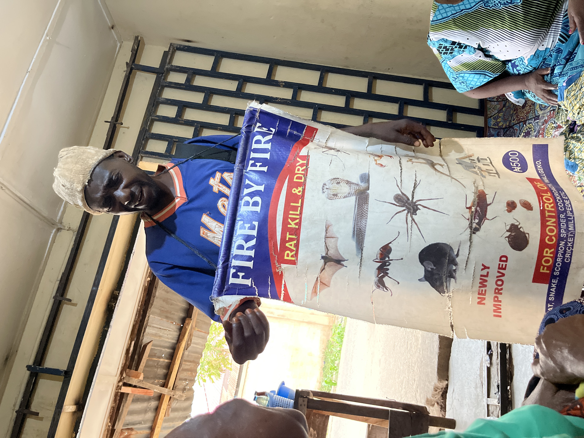 A man stands, holding a poster, titled "FIRE BY FIRE," advertising pest control. The poster displays images of rats, snakes, scorpions, spiders, cockroaches, wall geckos, crickets, and millipedes. The poster states it is newly improved and sells for ₦500. The location is Jos, Nigeria, as indicated by the currency symbol.
