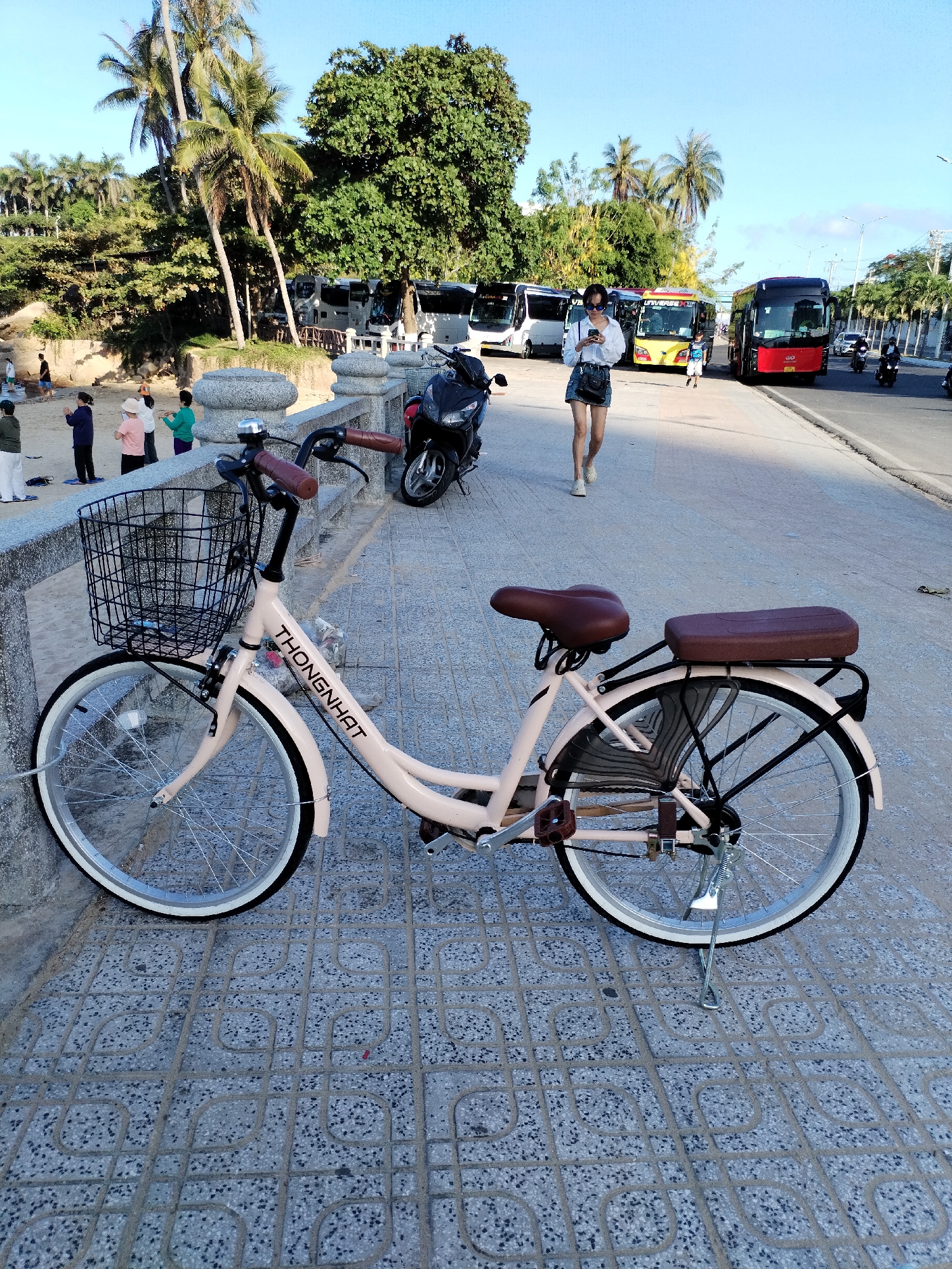 A light-pink Thong Nhat bicycle with a wire basket and brown seat is parked on a gray-tiled walkway. A woman wearing a white shirt and blue shorts walks on the sidewalk in the same direction as the parked bicycle in Bac Nha Trang Ward, Vietnam. Behind her are several buses and parked motorcycles. In the distance, trees, including palm trees, and people can be seen on a sandy beach. Other pedestrians are also walking on the roadway.