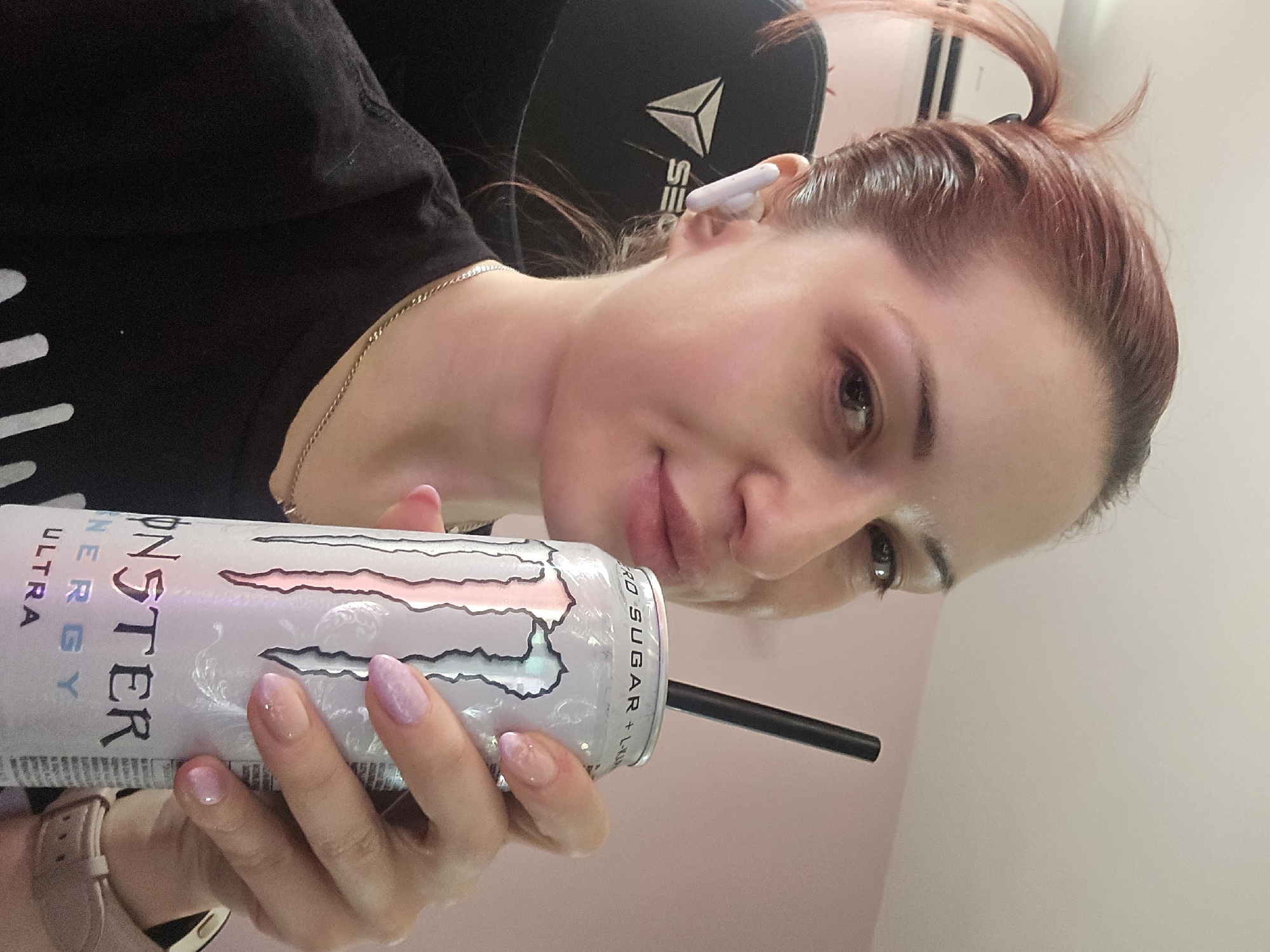 The image shows a woman holding a can of Monster Energy drink, with a black straw inserted. She has red hair pulled back in a ponytail and is wearing a black t-shirt and a silver chain necklace. She also has white earbuds in her ears. The background appears to be a room with a pink wall and a black gaming chair with a white logo. The woman is looking towards the camera and smiling slightly. There are no indications of the city of Burgas, Bulgaria within the image.