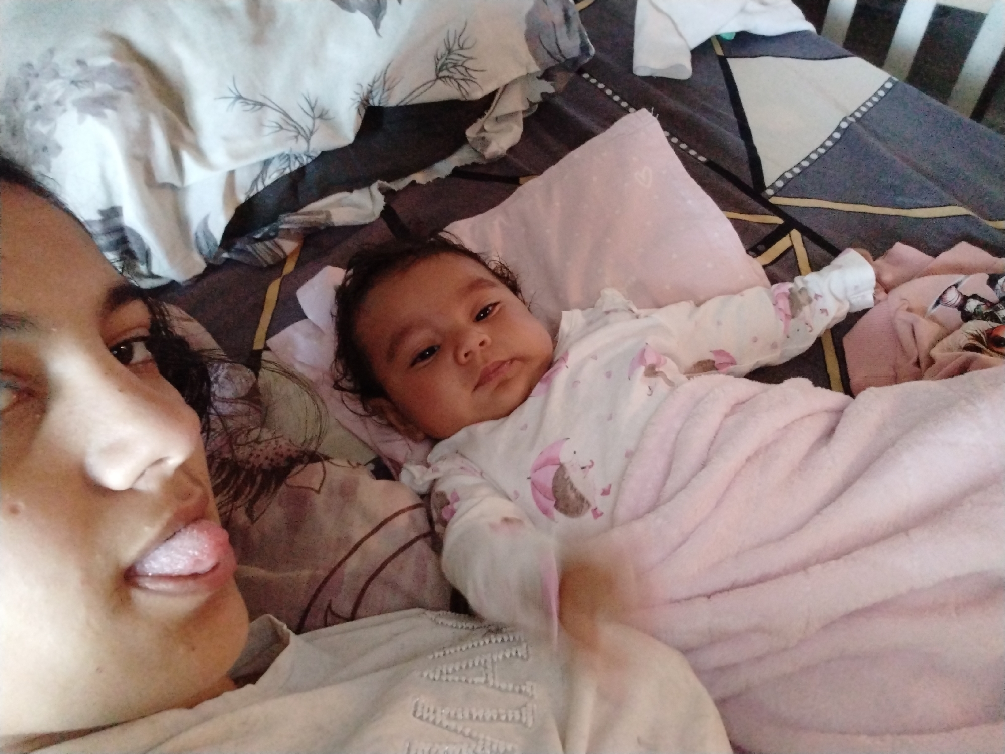 A selfie shows a woman with her tongue extended, lying next to a baby. The baby is lying on a bed, covered with a light pink blanket, and resting its head on a small pink pillow. The woman is wearing a white shirt and the baby is wearing a white onesie with a hedgehog print. The bed has patterned sheets and multiple pillows. The location is unknown.