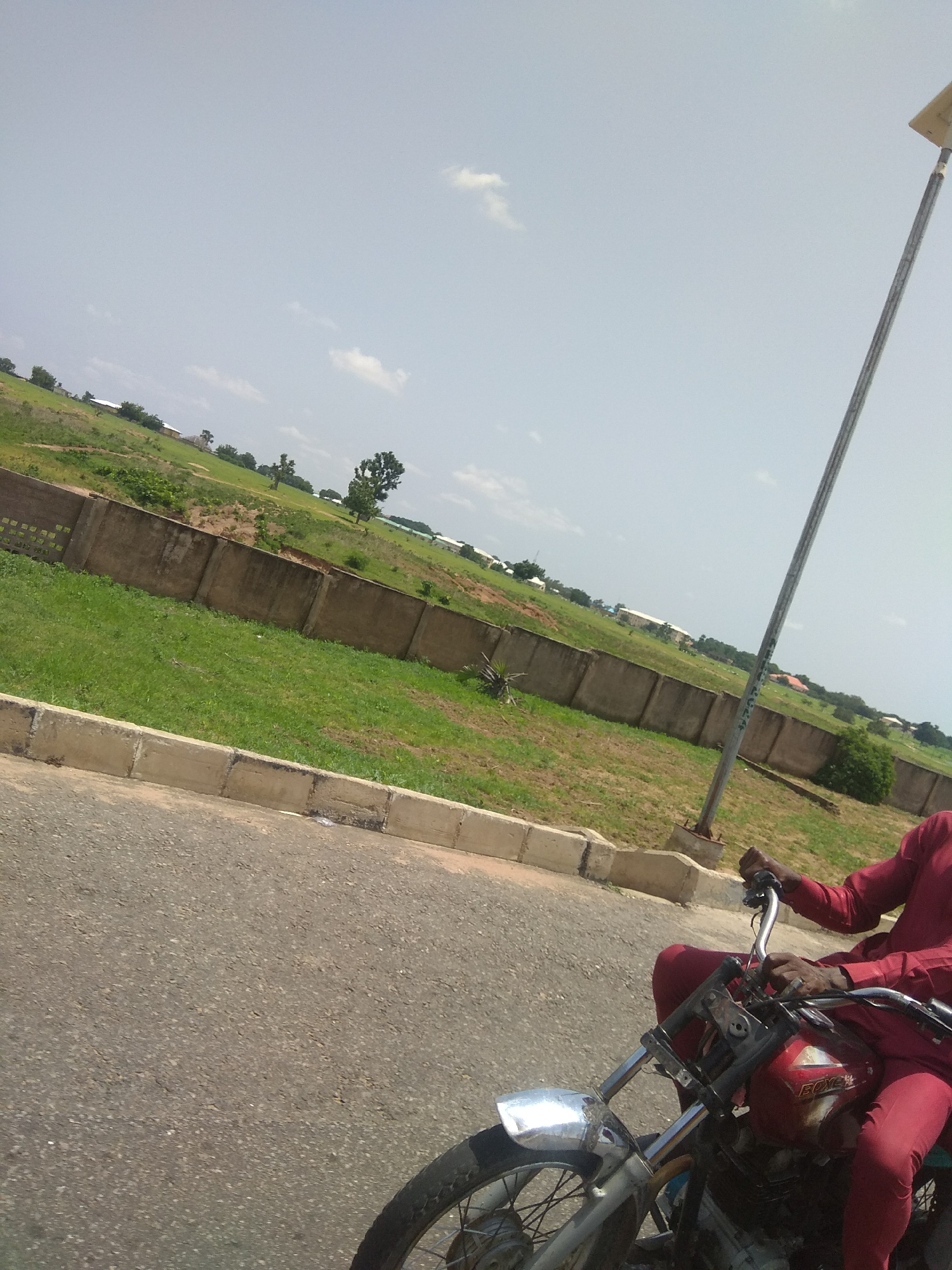 A person wearing a maroon outfit rides a motorcycle down an asphalt road. The motorcycle is red with some visible markings. A metal pole, likely a lamppost, stands to the right of the motorcycle. Behind the road is a grassy area bordered by a concrete wall. Beyond the wall, the landscape consists of more grassy fields, some scattered trees, and distant buildings. The sky is clear with a few wispy clouds. The scene appears to be located in a semi-urban or developing area of Jalingo, Nigeria.