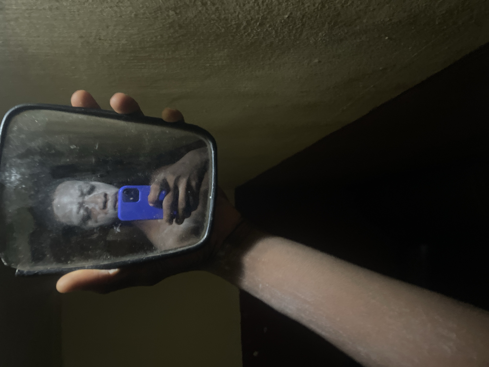 A close-up, angled photograph captures a person holding a rectangular mirror, reflecting their face and a smartphone. The mirror, held by a hand and forearm extending from the lower right of the frame, is dark-framed and appears smudged or dusty. Within the mirror's reflection, a person's face is visible, covered in a white substance, possibly a facial mask or makeup. Their hand, holding a bright blue-cased smartphone, is also reflected, indicating they are taking a picture of their reflection. The arm holding the mirror exhibits a somewhat dry or dusty texture.
The scene is dimly lit, suggesting an indoor setting, possibly at night or in a poorly lit room. Above the mirror and to the left, a textured, light-colored ceiling or upper wall is visible, while the area to the right is obscured by deep shadow, creating a strong diagonal line across the upper right quadrant of the image. The general atmosphere is subdued due to the low light. No specific location cues are visible within the image itself beyond the provided context of Onitsha, Nigeria. No text is discernible.