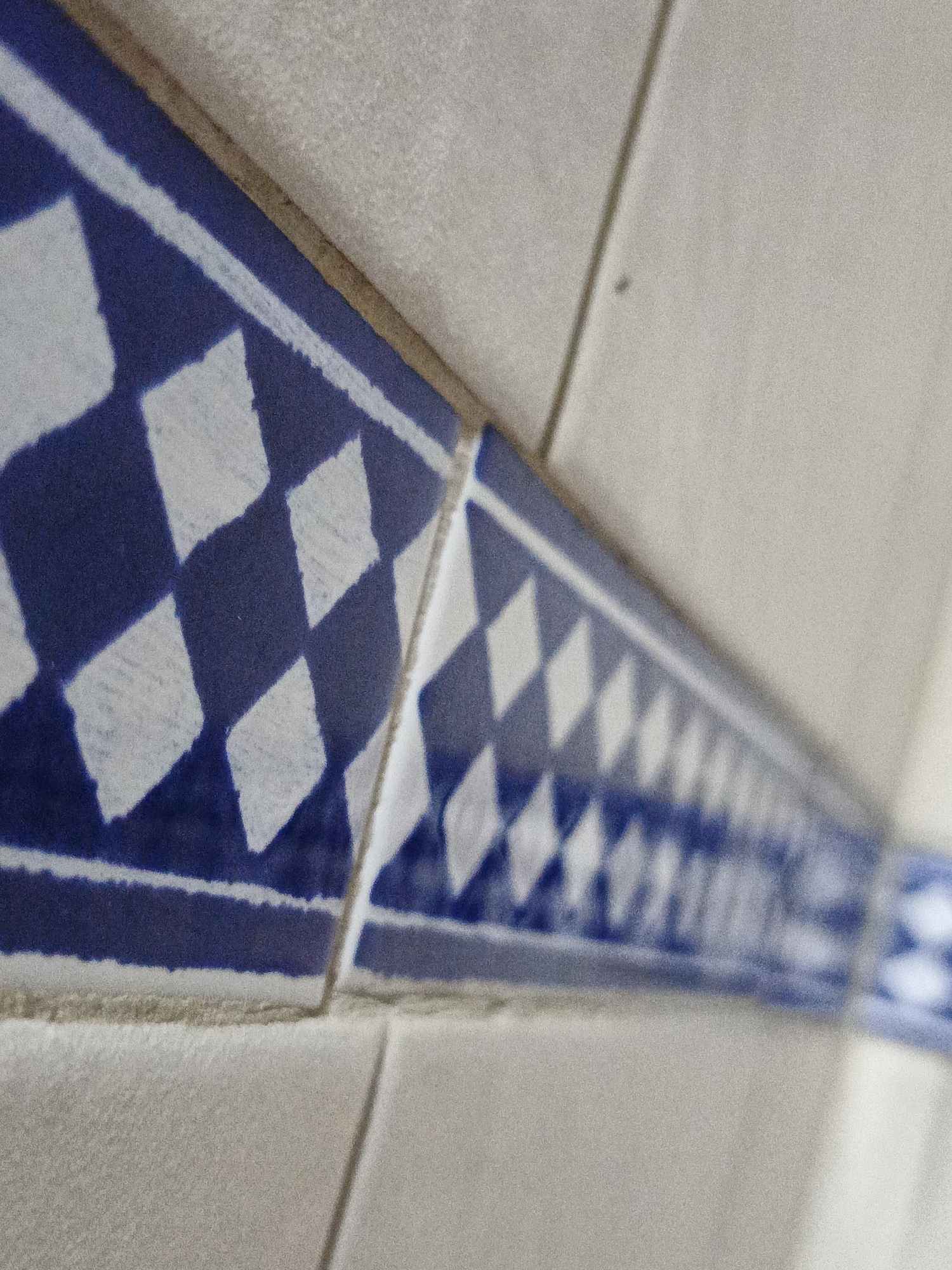 The image depicts a section of a tiled wall, likely indoors, with white tiles and a decorative border featuring a blue and white diamond pattern. The tiles are arranged in a linear fashion. The perspective is close-up and angled, causing a depth of field blur. The location in the city of Los Robles, Venezuela, is not evident.