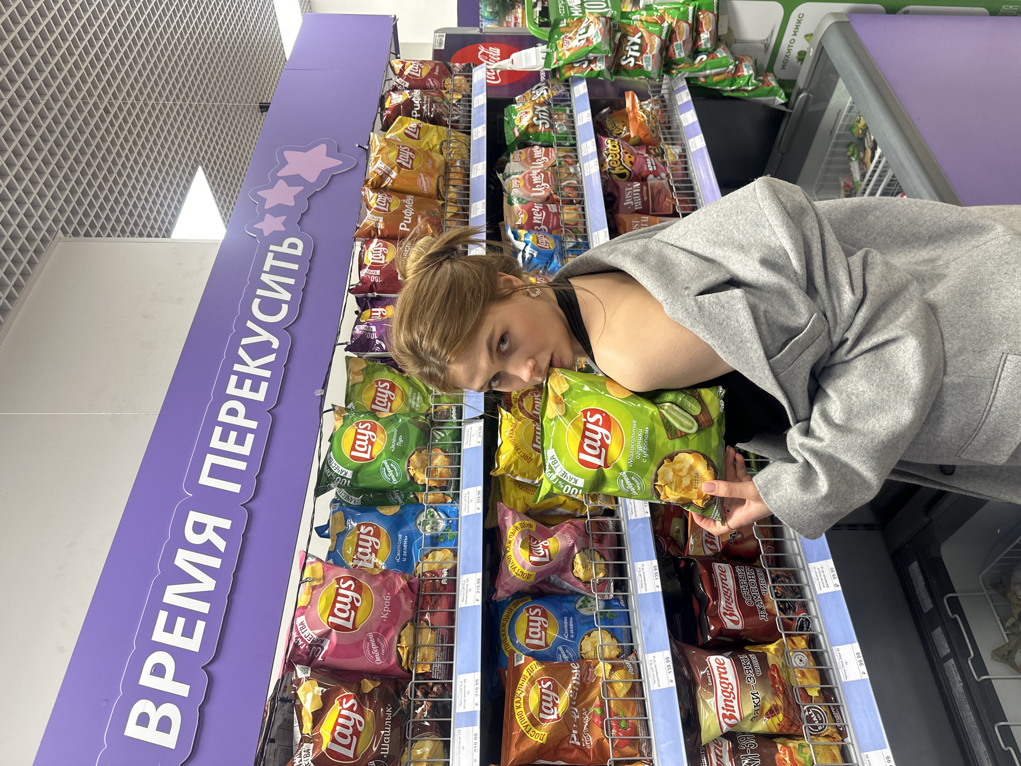 A young woman with blonde hair is selecting a bag of Lay's potato chips from a shelf in a grocery store in Krasnoyarsk, Russia. She is wearing a gray blazer over a black top. The shelves are stocked with various flavors of Lay's chips and other snacks. A sign above the chips reads "ВРЕМЯ ПЕРЕКУСИТЬ" in Russian, meaning "Time to Snack". There is a freezer unit visible to the right of the chip display.