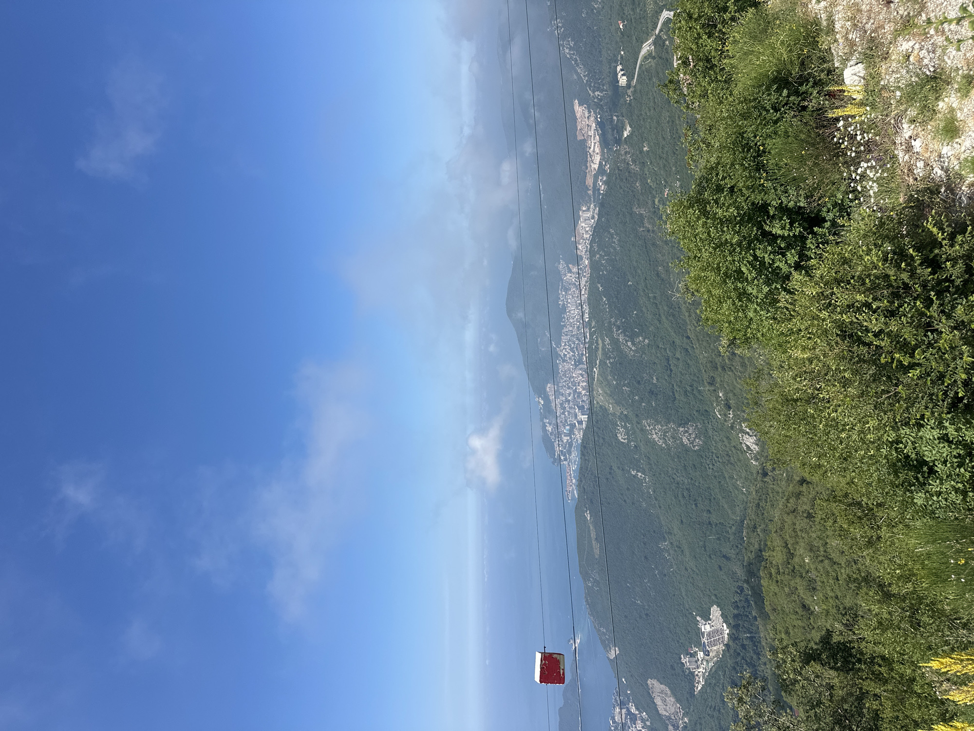 The image is an elevated, wide-angle shot of a coastal landscape with a city nestled between lush green mountains and the blue sea under a bright sky with scattered clouds. In the foreground, dense green bushes and some wildflowers are visible on a rocky incline. Several thin, dark cable car wires stretch across the frame, one of which holds a small, red and white cable car cabin. In the midground, a verdant, mountainous terrain slopes down to a city with numerous buildings, including some under construction, and a winding road. The city is situated along the coastline, with the vast expanse of the sea beyond. The background features distant, misty mountains and a clear blue sky. The scene appears to be taken from a high vantage point, likely a mountain overlooking the city of Brajici, Montenegro.