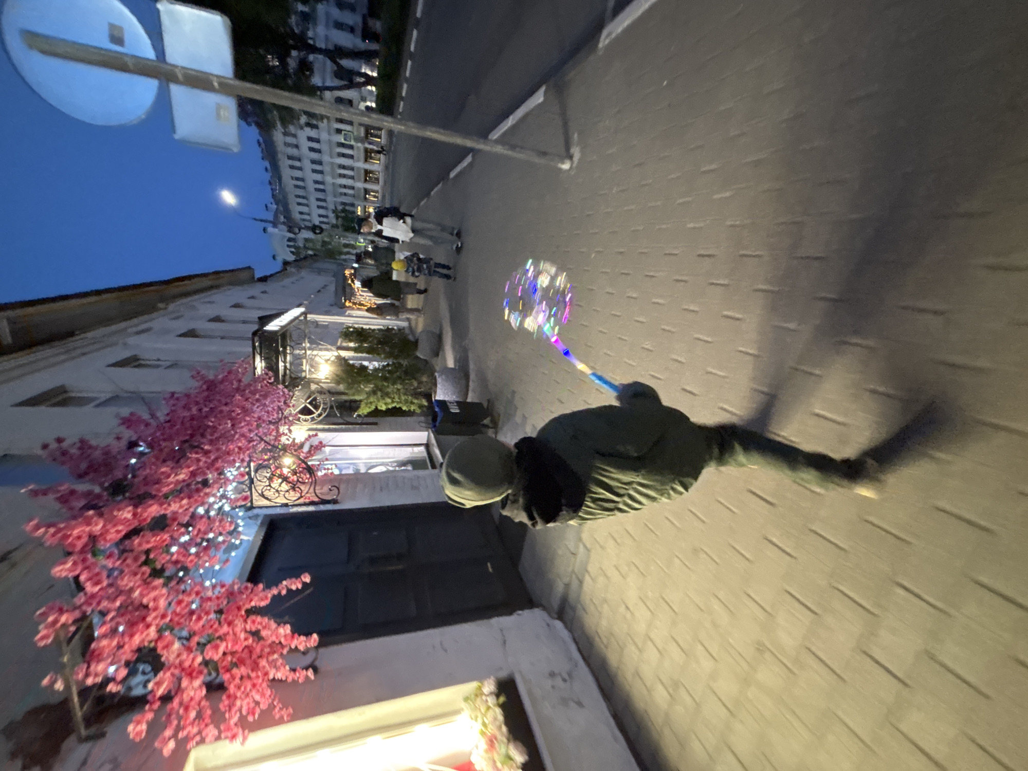 A person in a green jacket and gray cap walks down a sidewalk in Rusanovo, Russia, carrying a lit bubble wand. To the left is a building adorned with pink cherry blossom decorations and illuminated lights. Further up the sidewalk, other people are walking in the same direction. In the background, there is a multi-story building. Overhead is a street sign. The image was taken at dusk or night.