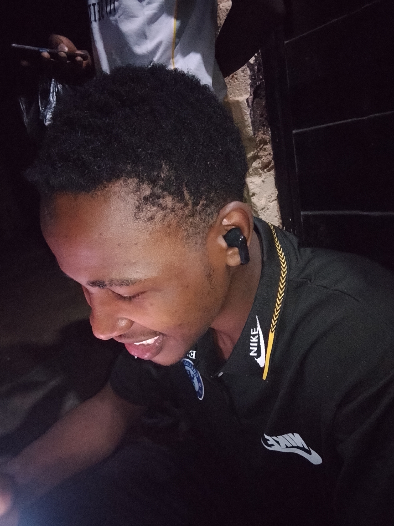 A close-up, outdoor, medium shot shows a young Black male from the shoulders up, looking down and to his left. He has short, dark, curly hair and is wearing a black polo shirt with a yellow and white patterned trim around the collar and a white Nike logo on the right sleeve. He has a black wireless earbud in his right ear and his mouth is open, revealing his teeth and tongue. The background is dark and out of focus, with hints of a textured, light-colored wall and a dark, ribbed structure. In the upper left corner, another person's hand is visible, holding a cell phone. The overall lighting is dim.