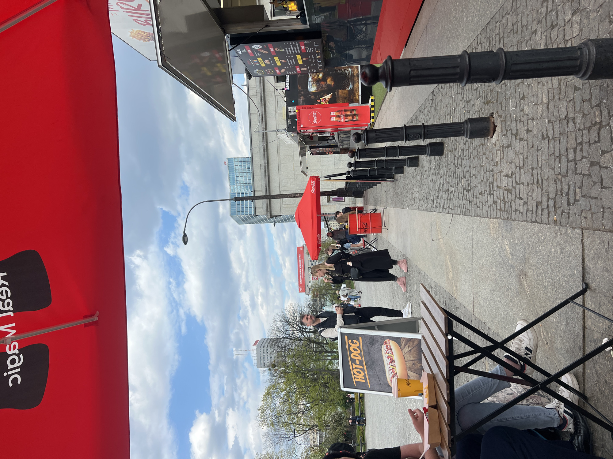 The image depicts a street scene in Warsaw, Poland, with an emphasis on a food vendor selling hot dogs. The photo is taken from the perspective of someone sitting at a table near the vendor's stall. A hot dog advertisement is visible on the table. People are walking along the sidewalk, some interacting with the vendor. There are red Coca-Cola umbrellas and branding. There are black bollards lining the walkway. The sky is partly cloudy, and the background includes buildings typical of an urban area.
