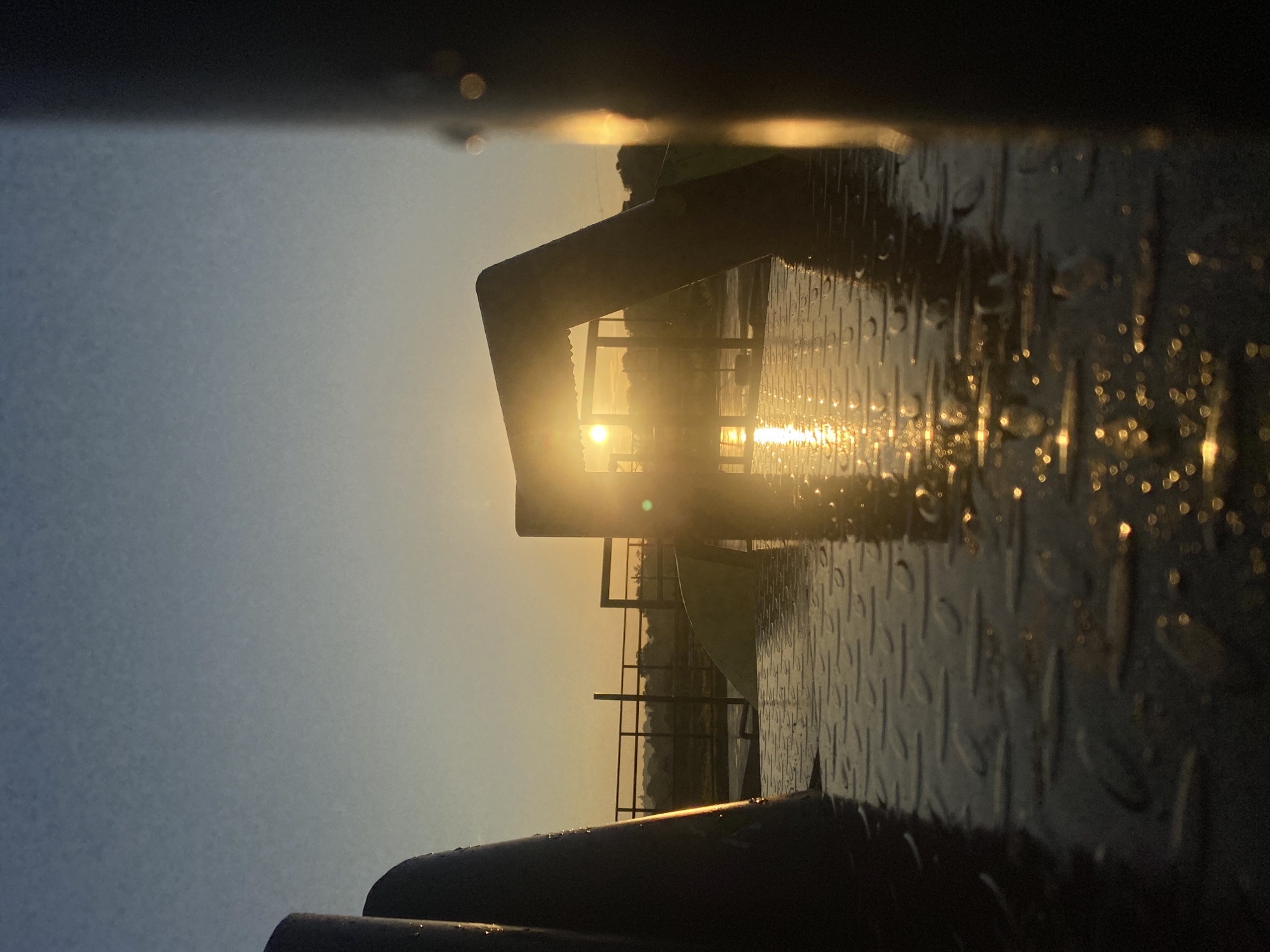 The image depicts a sunrise viewed from a low-angle perspective, likely on a platform or walkway. The foreground is dominated by a wet, textured metal surface, possibly diamond plate, reflecting the warm glow of the rising sun. Water droplets are visible on this surface, creating small, circular highlights. In the mid-ground, a metallic structure, possibly part of a playground or an outdoor amenity, partially obscures the sun. The sun itself is a bright orb, positioned in the center of the frame, with rays extending outwards and casting a strong lens flare. Beyond this structure, a railing runs horizontally, with a hazy landscape of trees and buildings visible in the distance, bathed in the golden light of dawn. The sky transitions from a clear, pale blue at the top to a warmer yellow-orange near the horizon. The scene suggests an outdoor location, potentially an urban park or waterfront area. Given the description of Davao City, this could be an area along the coast or a park with elevated structures offering sunrise views. There are no people visible in the image. The overall atmosphere is serene and tranquil, characterized by the soft light and reflections.