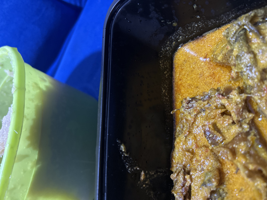 A close-up shot captured in Ilishan, Nigeria, displays two food containers on a blue fabric surface. In the foreground, a black rectangular plastic container holds a quantity of stew or soup. This dish features pieces of what appear to be meat or fish, immersed in a thick, reddish-orange, oily sauce. The sauce exhibits a slightly granular texture. Visible residue and droplets are present on the inner rim of the black container. In the upper background, partially obscured, is a light green, translucent bucket-like container. The top portion of this green container holds a light-colored, possibly granular or semi-solid substance, while the lower visible section contains a lighter-colored liquid. No individuals are present in the frame.