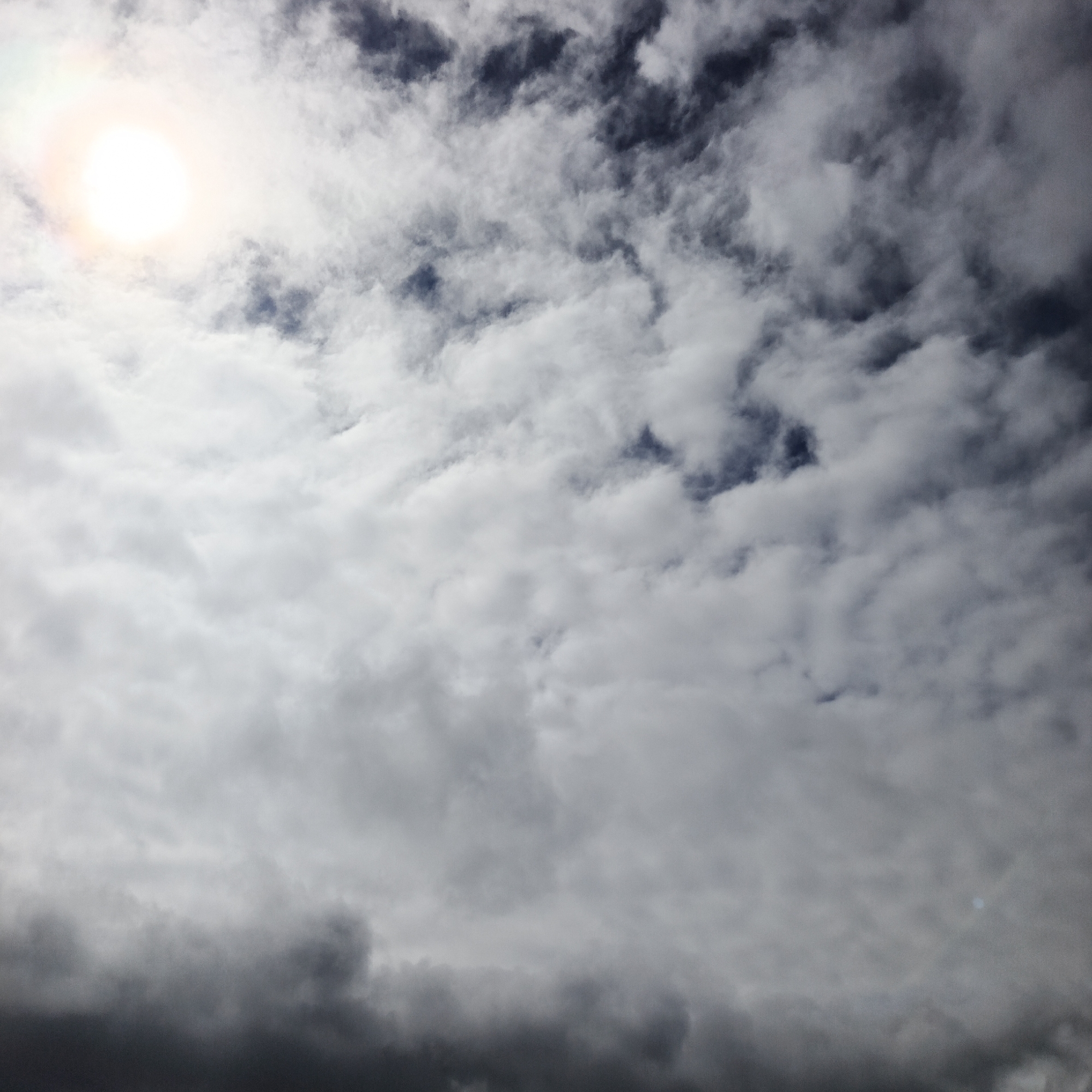 The image shows a cloudy sky with the sun visible in the upper left corner, creating a bright halo effect. The clouds are varied in density, with some appearing wispy and bright white, while others are darker and more ominous, suggesting potential rain or storm clouds. The overall scene depicts a dynamic sky. There are no people, subjects, or objects visible other than the sky and clouds. The location in Orlovka, Russia, cannot be determined from the visual information provided in the image.