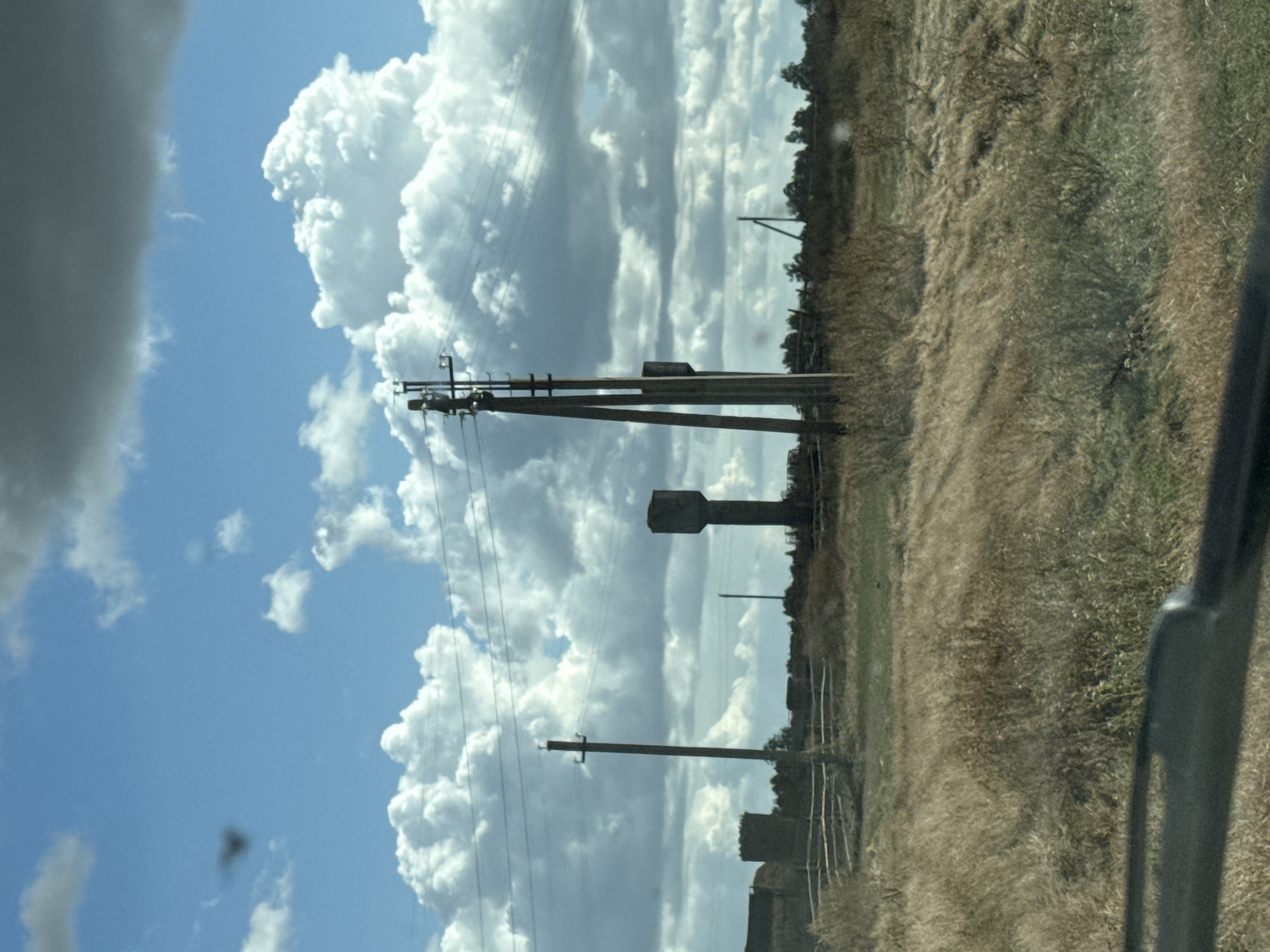 The image depicts a rural landscape under a cloudy sky. The foreground is dominated by dry, golden grass and low-lying shrubs, with a dark, blurry object, likely part of a vehicle's interior, visible at the bottom edge. In the mid-ground, a wooden fence runs horizontally across the frame. Behind the fence, two tall, concrete water towers stand prominently, one appearing slightly damaged. Several utility poles with power lines are also scattered throughout the scene. In the background, a line of trees forms a distant horizon. The sky is filled with large, fluffy white clouds interspersed with darker, gray clouds, suggesting an overcast or potentially stormy day. The scene is likely located in the vicinity of Fominka, Russia, given the architectural style of the water towers and the rural setting. No people are visible in the image.