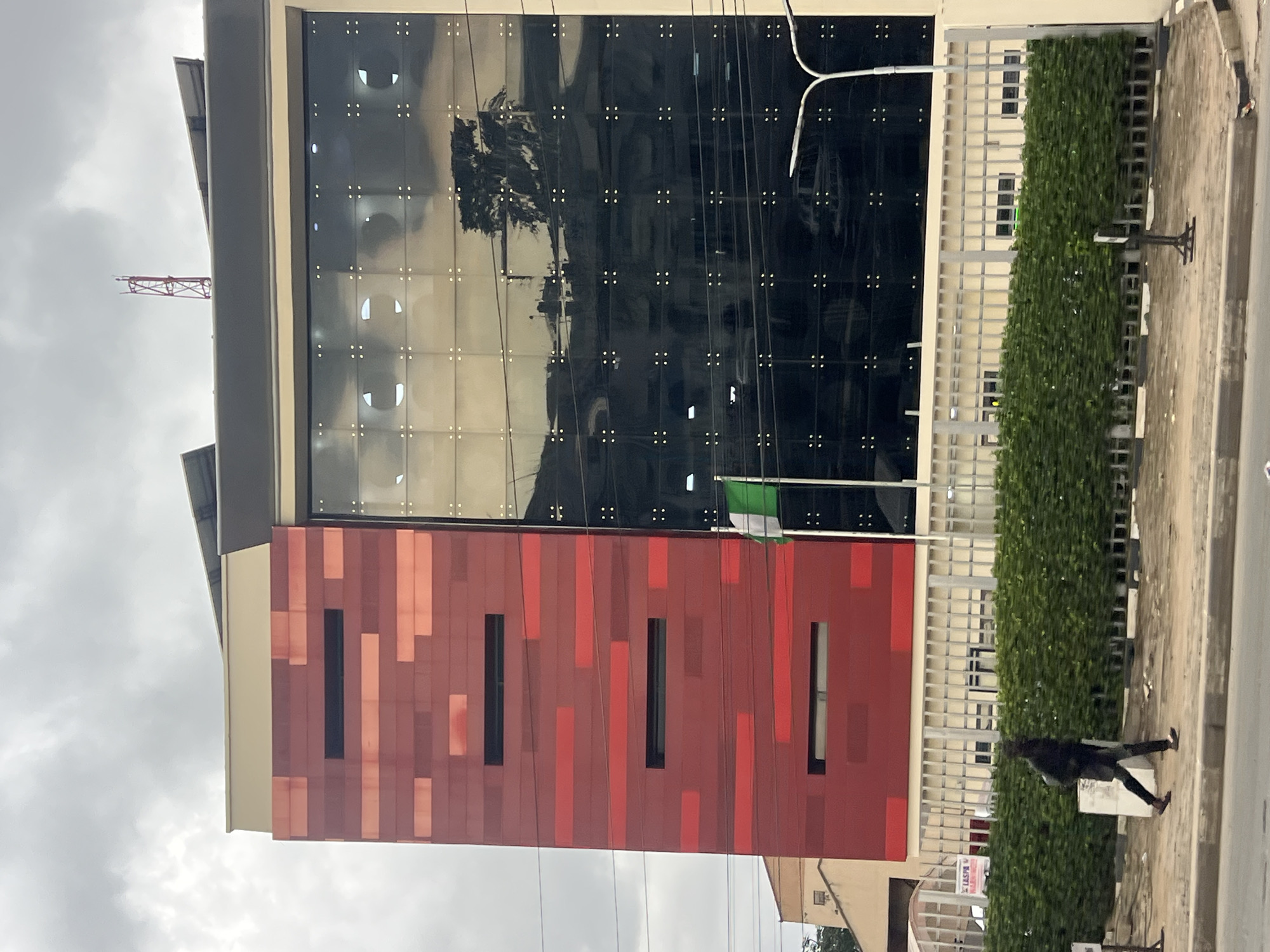 An overcast day in Ikeja, Nigeria, with a modern, multi-story building as the primary subject. The building features a striking facade with a section clad in alternating horizontal red and darker red rectangular panels, interspersed with narrow, dark rectangular windows. Adjacent to this is a large expanse of dark, reflective glass, mirroring the cloudy sky and surrounding trees. A Nigerian flag, green and white, flies from a pole near the glass section. Below the building is a low, light-colored fence backed by a dense, dark green hedge. A person wearing dark clothing, a backpack, and dark shoes is walking from left to right in front of the hedge. The ground in front of the hedge is a paved sidewalk and a dirt area with some debris. A street light with two lamps is visible to the right of the building, and a cell tower is visible on the roof.