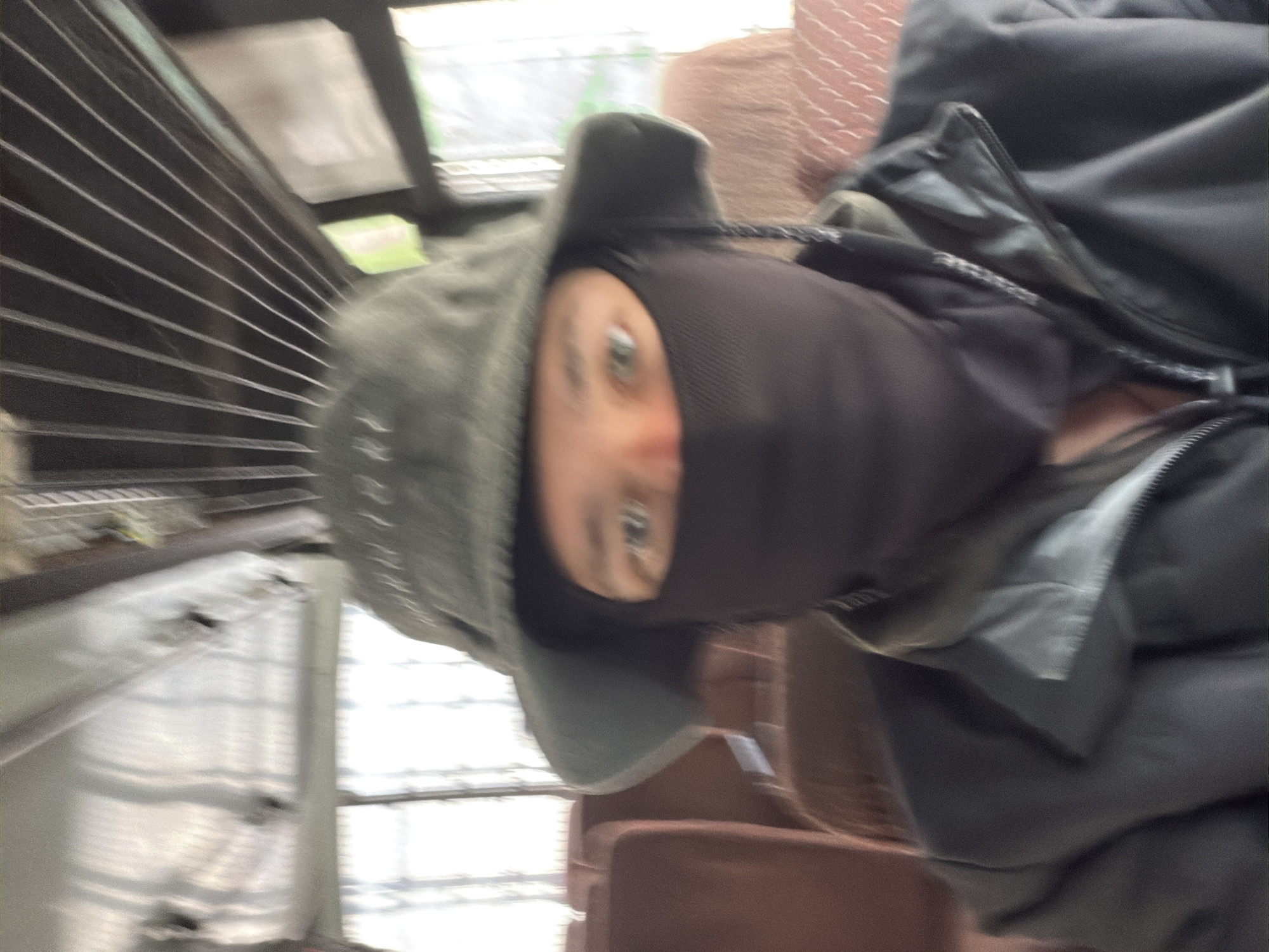 A blurred, first-person perspective photo shows a person wearing a dark balaclava and a green bucket hat. The person is sitting in a bus, with rows of brown seats visible behind them. The ceiling of the bus, with metal bars, is visible at the top of the frame. The photo appears to be taken in a city in India.