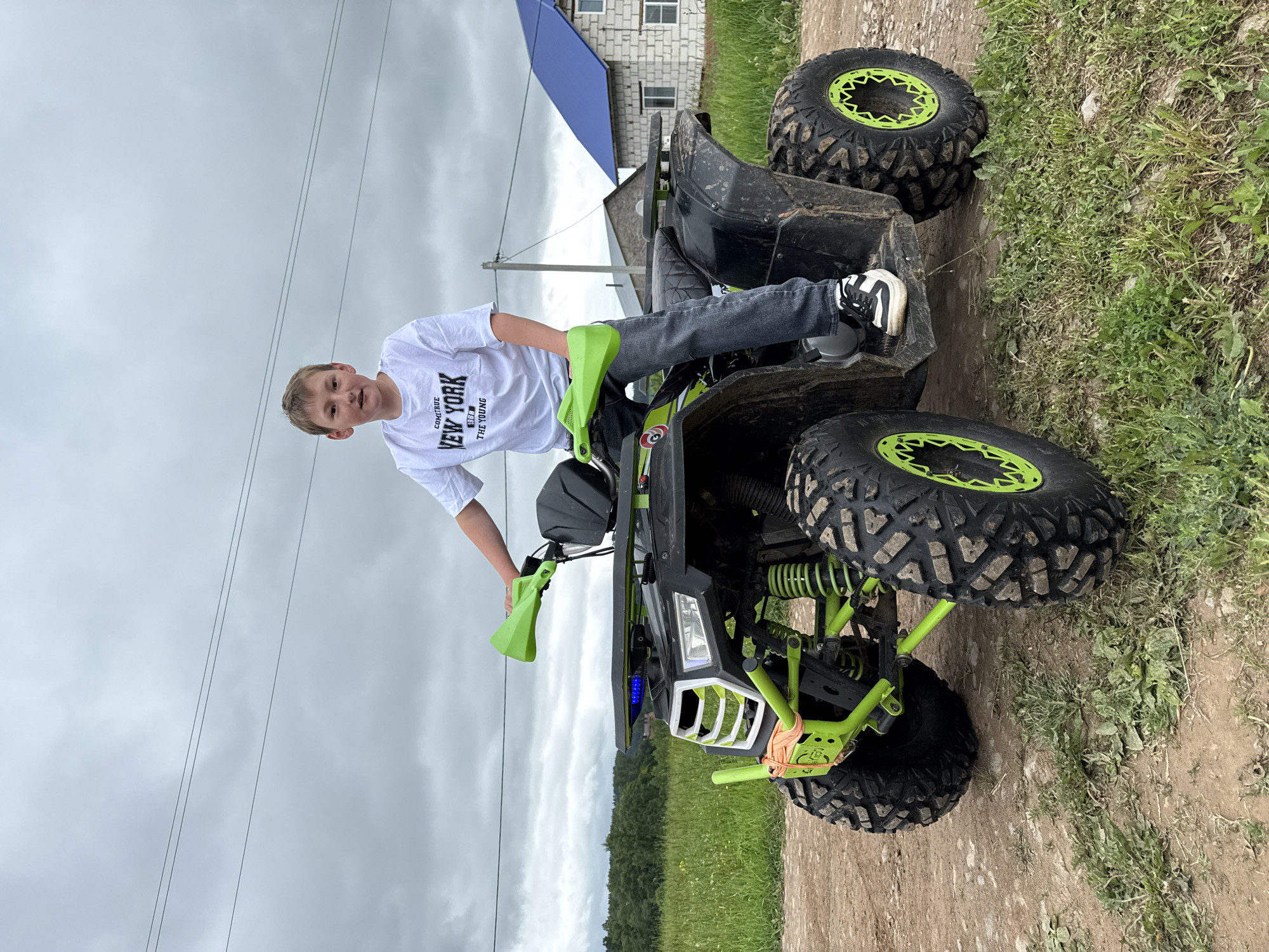 A young male, estimated to be between 8 and 12 years old, sits on a black all-terrain vehicle (ATV) with neon green accents, located on a dirt path in Ляльшур, Russia. The boy has short brown hair and fair skin, looking directly at the camera with a slight smile. He is wearing a white short-sleeved t-shirt with the text "COMITRUE NEW YORK 1968 THE YOUNG" printed in black on the chest, dark blue or black jeans, and black and white athletic shoes. His hands rest on the ATV's handlebars, which are fitted with neon green handguards.
The ATV features neon green front frame/bumper components, suspension springs, and wheel rims. Its body is black and appears soiled with dried mud. The front of the ATV includes a white grille, headlights emitting a bluish-white light, and a horizontal light bar above them displaying multiple blue LED lights. Large, knobby, muddy tires are fitted to the vehicle. An orange strap is visible secured around the front left suspension assembly.
The immediate foreground consists of a dirt path with patches of green grass and weeds. The background on the left features an open green field with tall grass, leading to a line of dense deciduous trees under an overcast sky filled with grey clouds. To the right, rural structures are visible, including a prominent white brick house with a distinctive bright blue corrugated metal roof and white-framed windows. A wooden utility pole with power lines spans the middle distance horizontally across the frame.