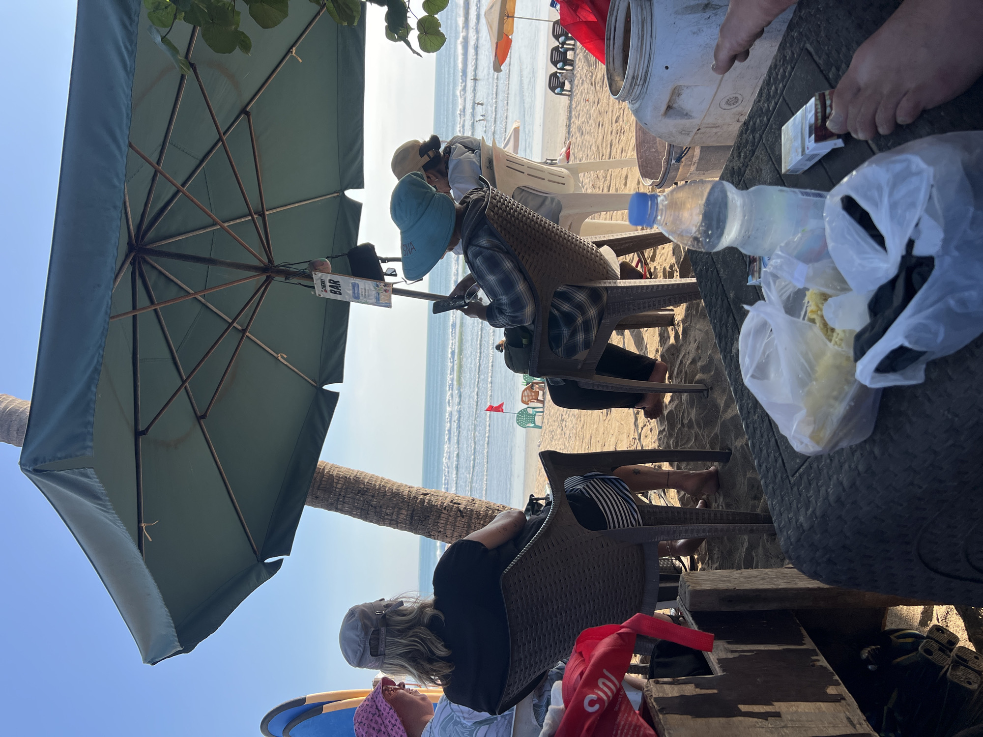 The image depicts a scene at a beachside establishment in Kuta, Indonesia. In the foreground, a dark-colored, textured table is laden with various items, including plastic bags containing food, a pack of cigarettes, a water bottle, and a large white plastic container. Several pairs of feet are visible, suggesting people are seated around the table. Beyond the table, three individuals are seated in chairs facing the ocean. The person on the left wears a pink headscarf. The central figure, facing away from the camera, has blonde hair, wears a grey cap, and a black t-shirt with striped shorts. To their right, another person, also facing away, wears a blue wide-brimmed hat and a plaid shirt, engrossed in their smartphone. In the background, the beach stretches out, with lounge chairs, umbrellas, and a few people in the water. A tall palm tree and a large green umbrella provide shade. The ocean has moderate waves, and the sky is clear and blue. A small red flag is planted on the beach. A sign hanging from the umbrella reads "CHERRY BAR." The overall atmosphere is relaxed and casual, indicative of a beach bar environment.