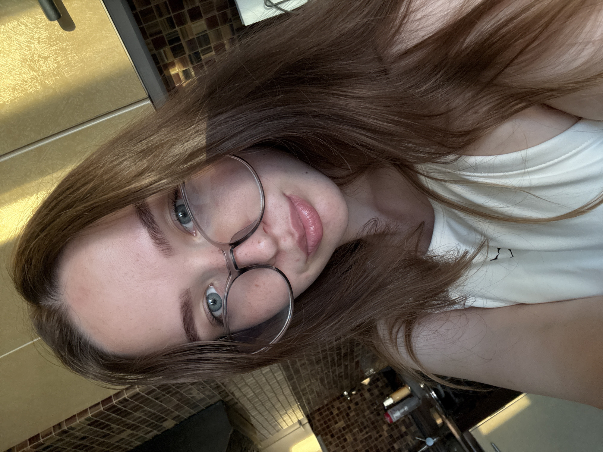 A selfie is taken from a high angle, showing a young person with long, light brown hair. They are wearing round, clear-framed glasses and a white t-shirt. Their eyes are blue-grey, and their lips are slightly parted, with a rosy hue. The background appears to be a kitchen, with tiled walls and a golden-colored surface that could be a countertop or appliance. The lighting suggests it is daytime, possibly late afternoon due to the warm tones. The person's expression is neutral or slightly curious. There is no visible text in the image. The scene is indoors and appears to be in a private residence.
