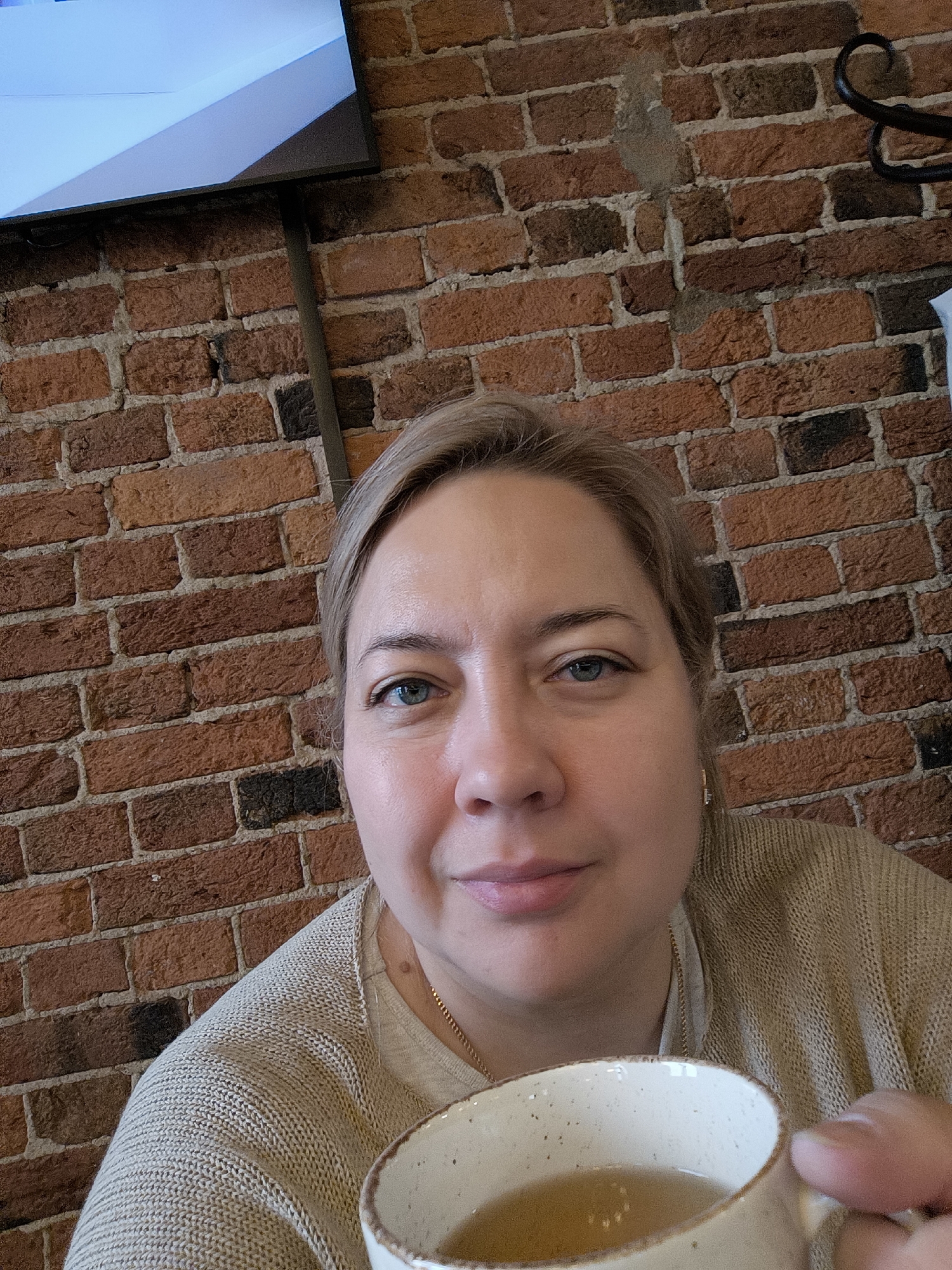 A woman is captured in a close-up selfie, holding a mug and looking directly at the camera with a slight, relaxed smile. She appears to be in an indoor setting, likely a cafe or restaurant, given the rustic brick wall background. The woman has light-colored hair, possibly blonde or light brown, pulled back or short, and blue eyes. Her skin has a natural, un-made-up appearance. She is wearing a light beige or cream-colored knit sweater over a lighter top, and a thin gold chain is visible around her neck.
In her right hand, she holds a ceramic mug with a speckled, off-white exterior and a brown rim, containing a light amber-colored liquid, likely tea or another warm beverage. The mug is positioned in the lower right corner of the frame.
The background is dominated by a textured brick wall with varying shades of red, orange, and brown bricks set with light-colored mortar. In the upper left corner, a portion of a modern, flat-screen television or monitor is visible, displaying a light blue or grey screen with a diagonal dark line. A dark, possibly metal, pole or support structure runs vertically from behind the screen down the brick wall. Another detail on the right side of the brick wall shows what looks like a black, decorative metal scrollwork, possibly part of a hanger or fixture.
The lighting appears soft and even, suggesting an indoor environment without harsh direct sunlight. The overall mood conveyed is calm and informal. No visible text is legible in the image. Given the location context, the scene is set in Moscow, Russia.