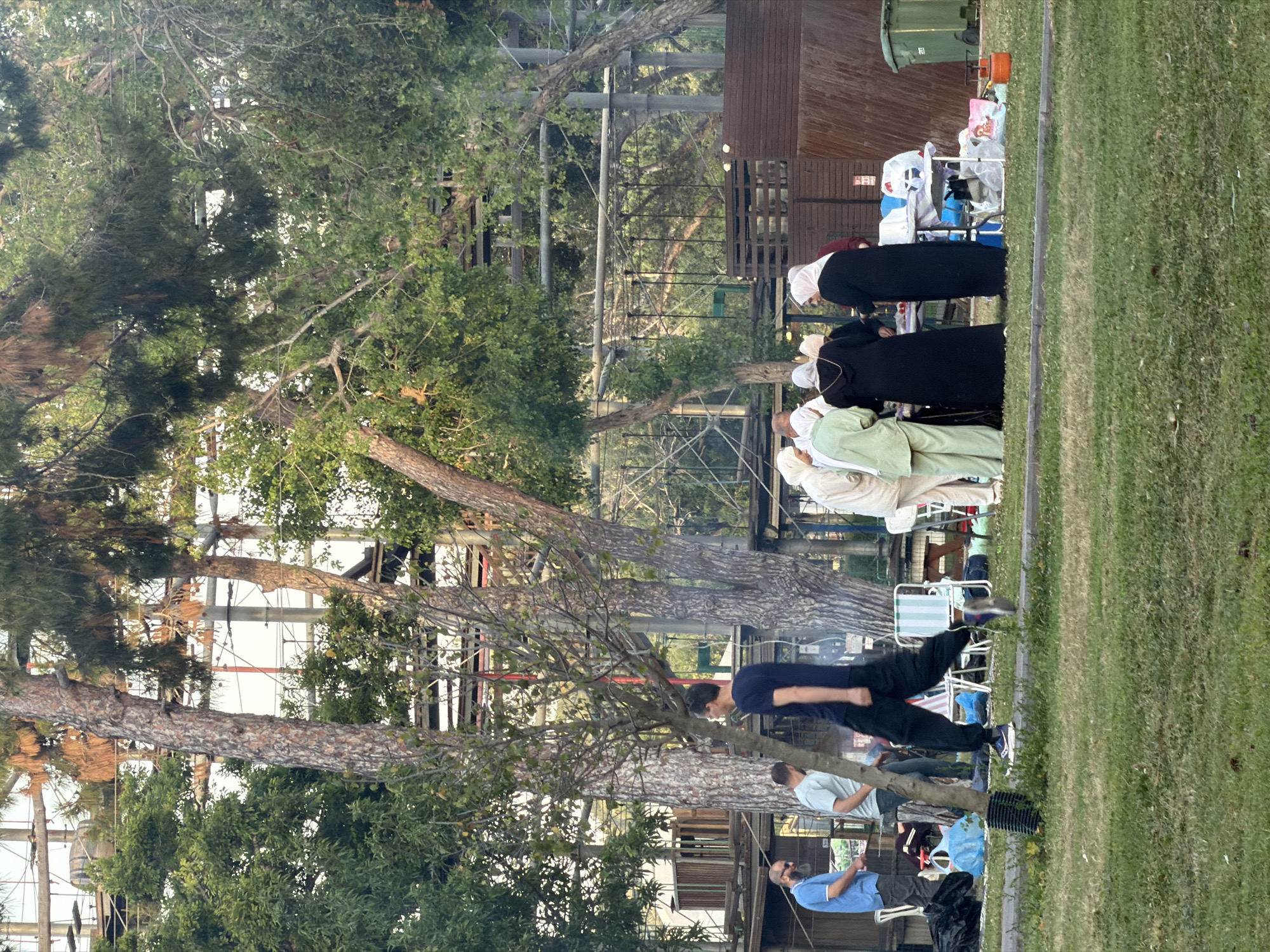 People are gathered outdoors in Tel Aviv, Israel. Several individuals wearing head coverings and long robes are standing in a line on a grassy area. Near them, a few other people are walking and standing. In the background, there are trees, scaffolding, and a building. The scene appears to be a public space, possibly a park.