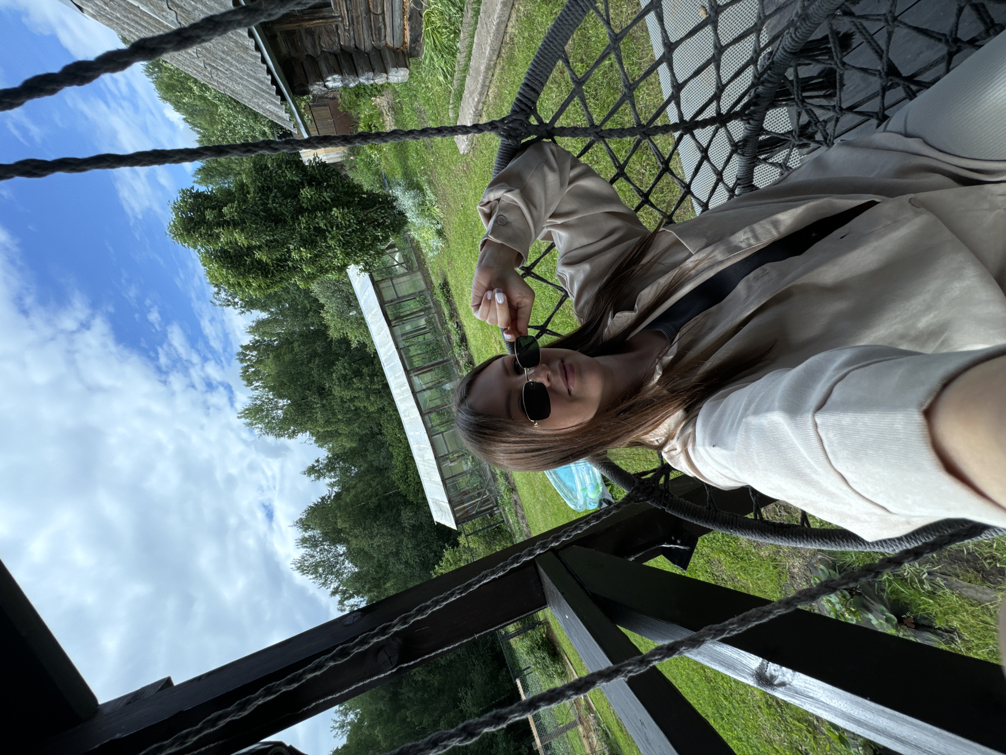 A young woman with long brown hair and sunglasses sits in a netted swing chair outdoors, on a wooden porch overlooking a rural landscape. She is wearing a light beige shirt and pants. In the background, there is a greenhouse, a wooden log cabin, trees, and a blue inflatable pool. The sky is partly cloudy. The location is the village of Ponaryevo, in the Podolsky District of Moscow Oblast, Russia.