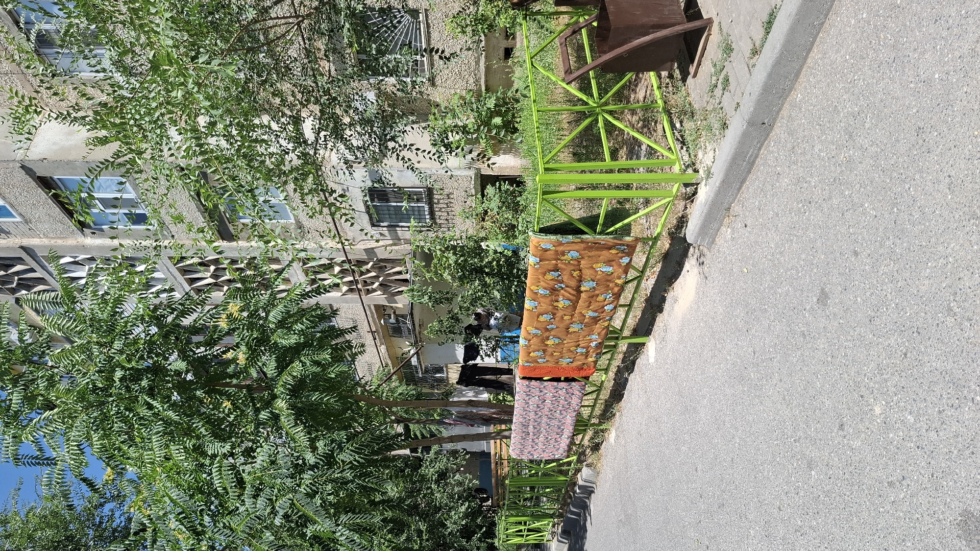 In Shymkent, Kazakhstan, a sunny day is captured on a street with an apartment building in the background and a bright green metal fence in the foreground. Two patterned blankets, one with a floral design and the other with a yellow and blue pattern, are hung on the fence to dry. The apartment building has a rough, concrete facade with geometric window frames and a balcony. Lush green trees and bushes surround the building and the fence, indicating a well-treed urban environment. The street is paved with asphalt, and a concrete curb runs alongside the sidewalk. To the right of the fence, a brown metal trash bin is visible. The scene suggests a typical residential area in Shymkent, where daily life activities like drying laundry outdoors are common.