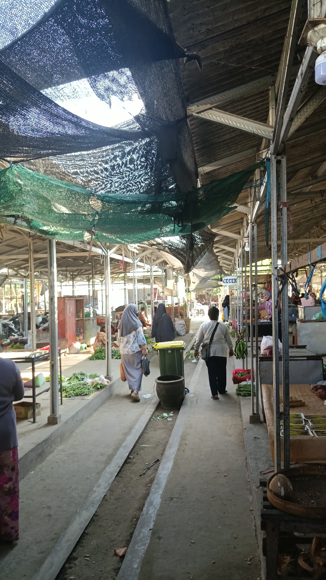 The image depicts an outdoor marketplace with a covered walkway in Purwokerto, Indonesia. The walkway is constructed with metal poles and a corrugated metal roof, partially covered by dark netting and green mesh, creating shade. In the foreground, a person in a purple and pink patterned skirt is visible from the waist down, standing on the left. Further into the market, a woman in a grey hijab and a floral patterned top walks away from the camera, carrying a dark bag. Behind her, another person in a black hijab is also walking. To the right of these individuals is a large, dark green trash can. Along the walkway, piles of fresh green vegetables are displayed on the ground and on low tables. In the mid-ground, several stalls are visible, with people interacting and selling goods. A sign reading "CECEK" is visible above one of the stalls. The ground of the walkway is concrete with a drainage channel running down the center. In the background, more stalls and people can be seen, indicating a bustling market environment. The overall lighting suggests it is daytime.