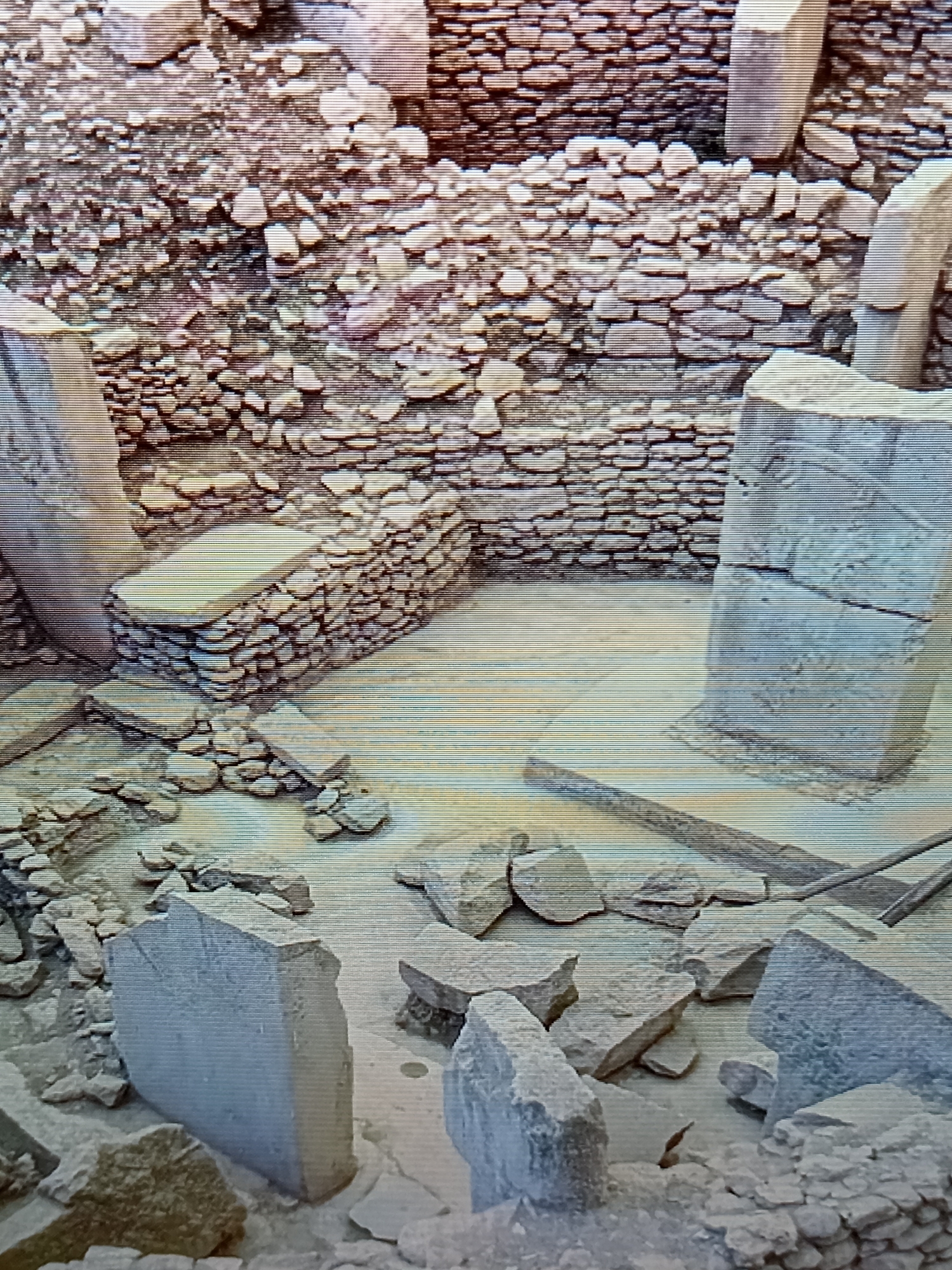 The image depicts an archaeological excavation site. In the foreground, several large, irregularly shaped stone blocks are scattered across a dusty ground. Some of these blocks are upright, while others are lying flat or broken. Behind these, a paved area is visible, with a low, circular stone structure or platform in the center. To the left, a tiered stone platform or seating area is constructed from smaller stones. Further back, a wall made of rough-hewn stones forms the backdrop of the scene. The overall impression is of an ancient ruin being uncovered. This site is not located in Brno, Czechia.