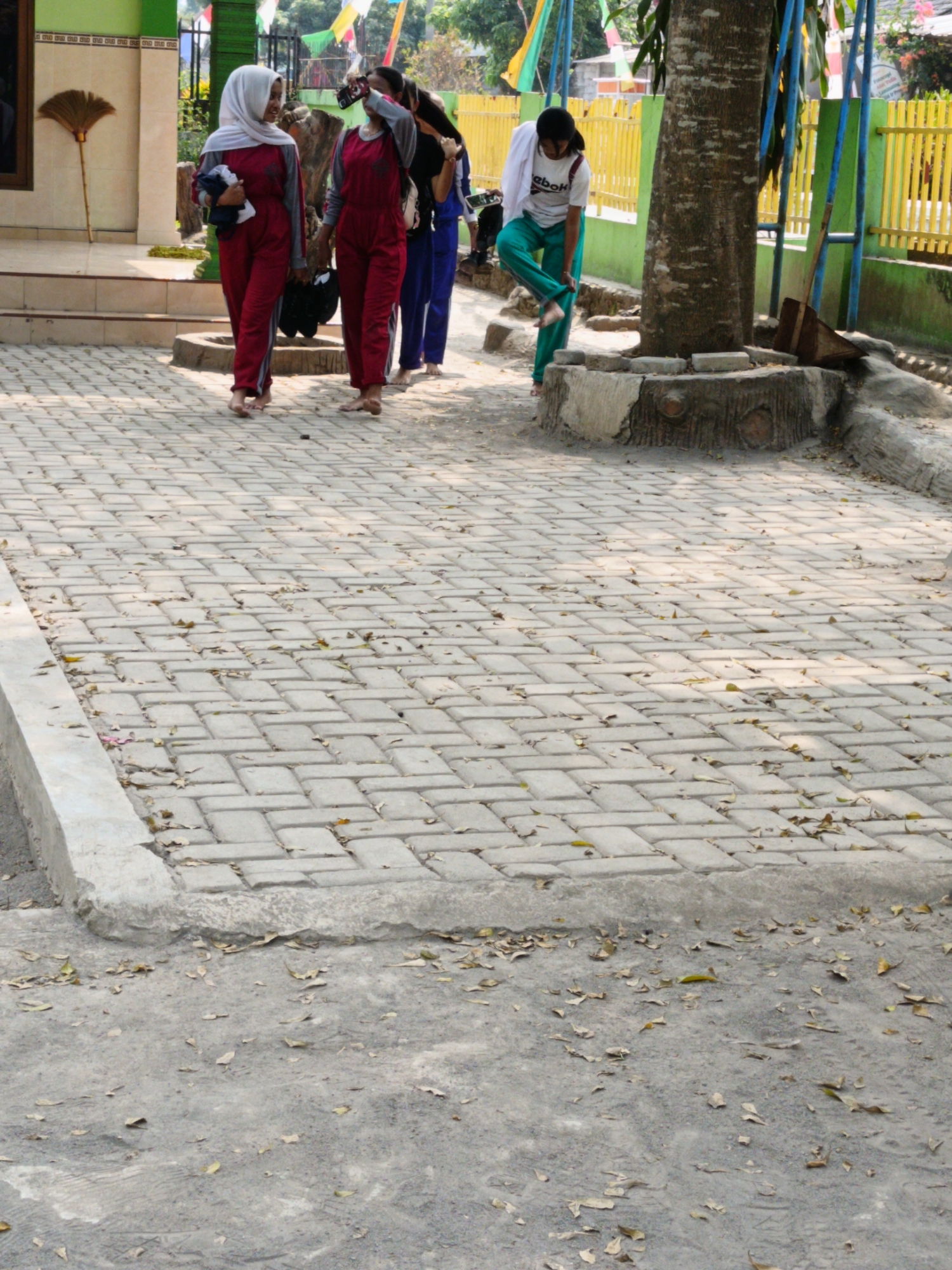 In Barukan, Indonesia, a group of four individuals is walking on a paved walkway. The walkway is made of light-colored interlocking bricks, with fallen leaves scattered across its surface. A low concrete curb lines the edge of the walkway.
The first person, on the left, is a woman wearing a white hijab and a dark red long-sleeved shirt and matching pants. She is barefoot and holding a dark-colored bundle in her left arm.
Next to her is another woman, also barefoot, wearing a dark red long-sleeved shirt and pants, similar to the first woman.
To their right, a third individual, a woman, is wearing a white t-shirt with "Reebok" printed on it, and teal-colored pants. She is barefoot and is in the process of adjusting her pants.
The fourth individual, furthest to the right, is partially visible and appears to be wearing dark clothing.
In the background, a tree with a thick trunk stands near the walkway. A portion of a yellow metal fence is visible behind the tree. To the left of the walkway, a building with a green facade and a brown door is present. A broom with a straw head is leaning against the building. The scene appears to be outdoors, likely in a public area.