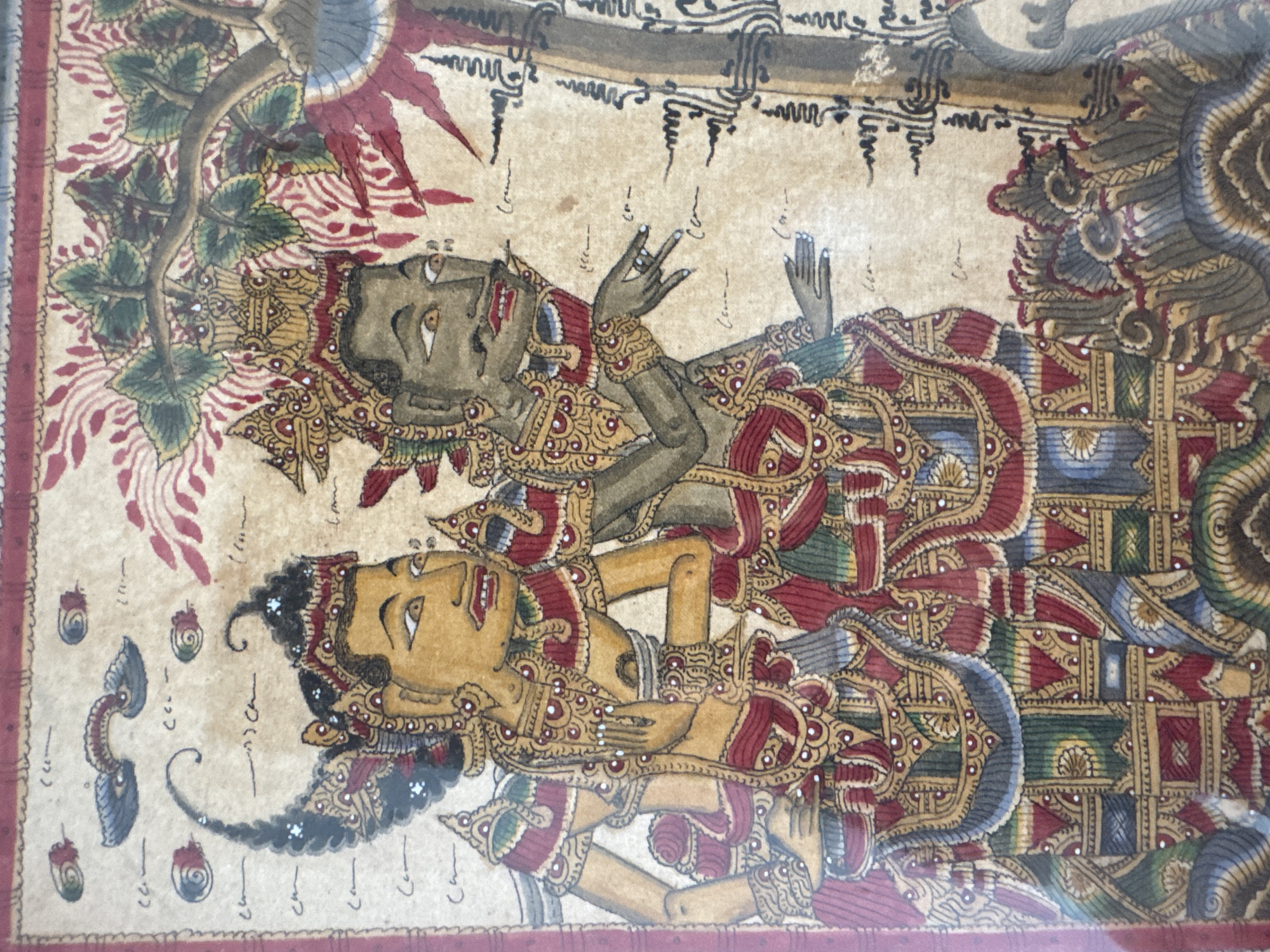 This is a close-up, eye-level shot of a Balinese painting depicting two figures in traditional attire, possibly Wayang Kulit or a similar form of shadow puppetry or illustration. The figures are rendered with a muted color palette dominated by earthy tones, reds, and golds, on a weathered, parchment-like background. Both figures wear elaborate headdresses and ornate, layered garments adorned with intricate patterns and beadwork. The figure on the left has yellowish skin and a stylized, elongated face with large, prominent eyes and lips. The figure on the right has darker, grayish skin, also with stylized facial features. Their hands are positioned as if in mid-gesture, with the figure on the right extending a finger. Behind and to the right of the figures, elements of a stylized tree with green leaves and red accents are visible, along with abstract patterns and possibly decorative borders along the top and left edges of the painting. The overall impression is one of ancient storytelling or performance art. The image is a photograph of the painting itself, not the scene it depicts, and therefore does not provide information about the city of Kuta Selatan, Indonesia.