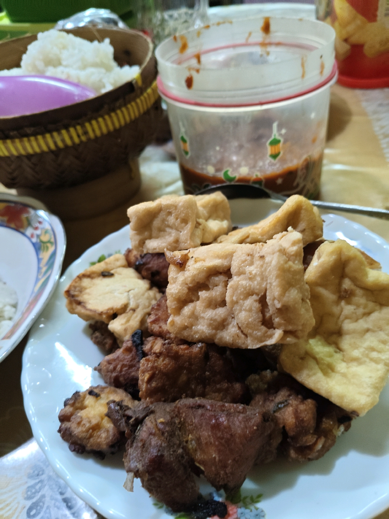 In the city of Sumberwono, Indonesia, on a table covered with a textured tablecloth, a white plate is filled with fried food, including pieces of fried tofu and meat. Beside the plate, a woven basket contains white rice, and a purple utensil rests on top of it. A clear plastic container, partially filled with a dark sauce, sits in the background, adorned with small decorative stickers of a lantern and crescent moon. The scene appears to be a casual meal setting.