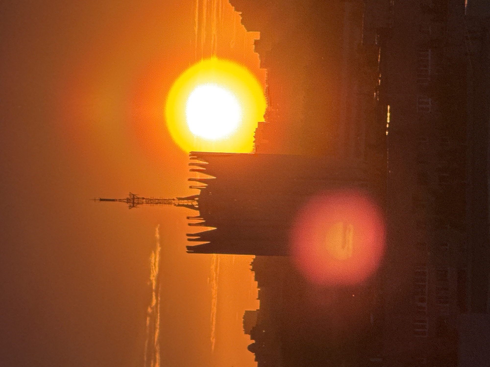 The image shows an orange sunset with a building in silhouette. A communications tower is atop the building. The location is likely an urban area. Another building can be seen to the right. The image also shows lens flare.
