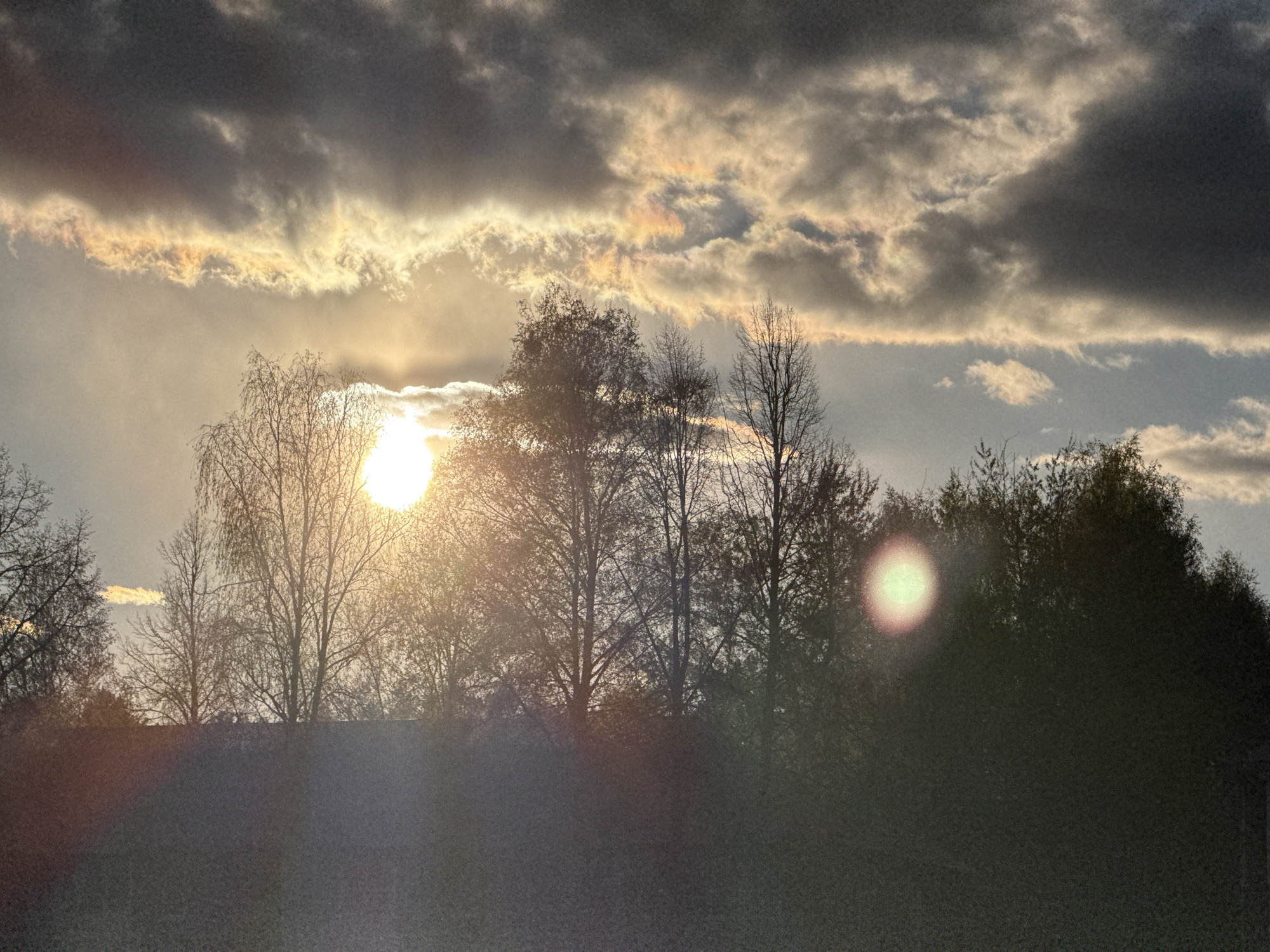 The image depicts a landscape with trees in the foreground silhouetted against a bright sky with the sun obscured by clouds. The sun's rays are visible, creating a lens flare effect. Some clouds show iridescence. The location is Votkinsk, Russia. There are no visible people or interactions.