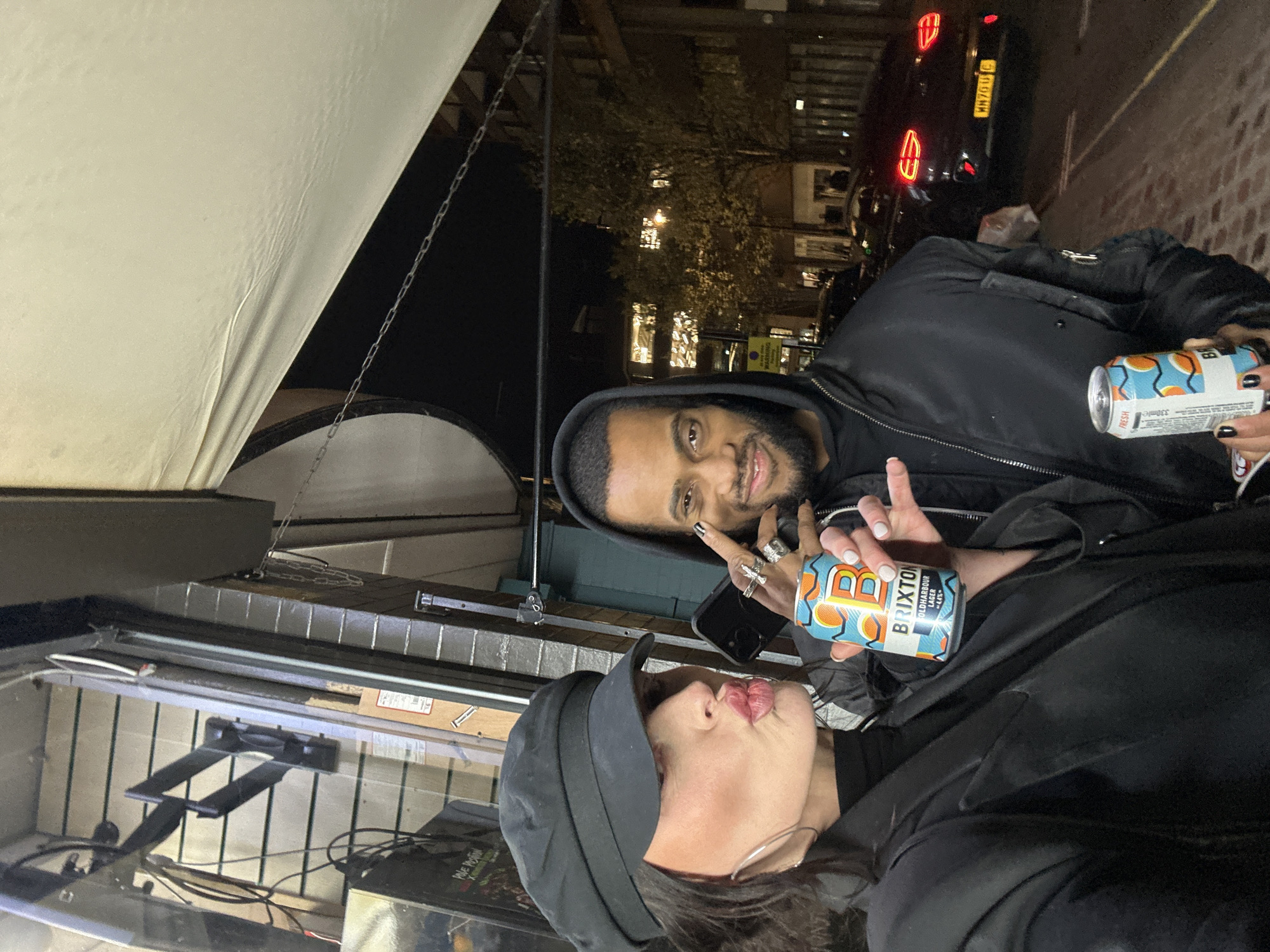 Two individuals are captured in a nighttime outdoor selfie, likely taken in Brixton, London, United Kingdom. The foreground shows a paved surface, possibly a sidewalk, with a light-colored awning or canopy extending overhead, secured by chains.
On the left, an adult female with dark hair wears a black newsboy-style cap, a black turtleneck, and a black blazer. Her face is turned slightly left, with lips pursed in a pout. She holds a blue and orange 'Brixton Oldharbour Lager 4.4%' can in her right hand. Her left hand, with white-painted fingernails, holds a black smartphone, suggesting she is operating the camera. Large hoop earrings are visible on her left ear.
On the right, an adult male with a beard and dark hair wears a dark hooded jacket. He smiles directly at the camera. His left hand, adorned with multiple rings and black-painted fingernails, is raised near his face. He holds an orange and white 'Brixton Fresh' can in his right hand.
The background reveals an urban street scene. To the immediate left of the female, a shop window displays internal fixtures, including slatwall paneling and electrical wiring. Behind the individuals, a street is visible with parked vehicles. A dark-colored car with distinct taillights displays a UK license plate "MN70 ULC". In the distance, multiple-story buildings with illuminated windows and trees are present under a dark sky. A yellow parking sign is partially visible behind the male.