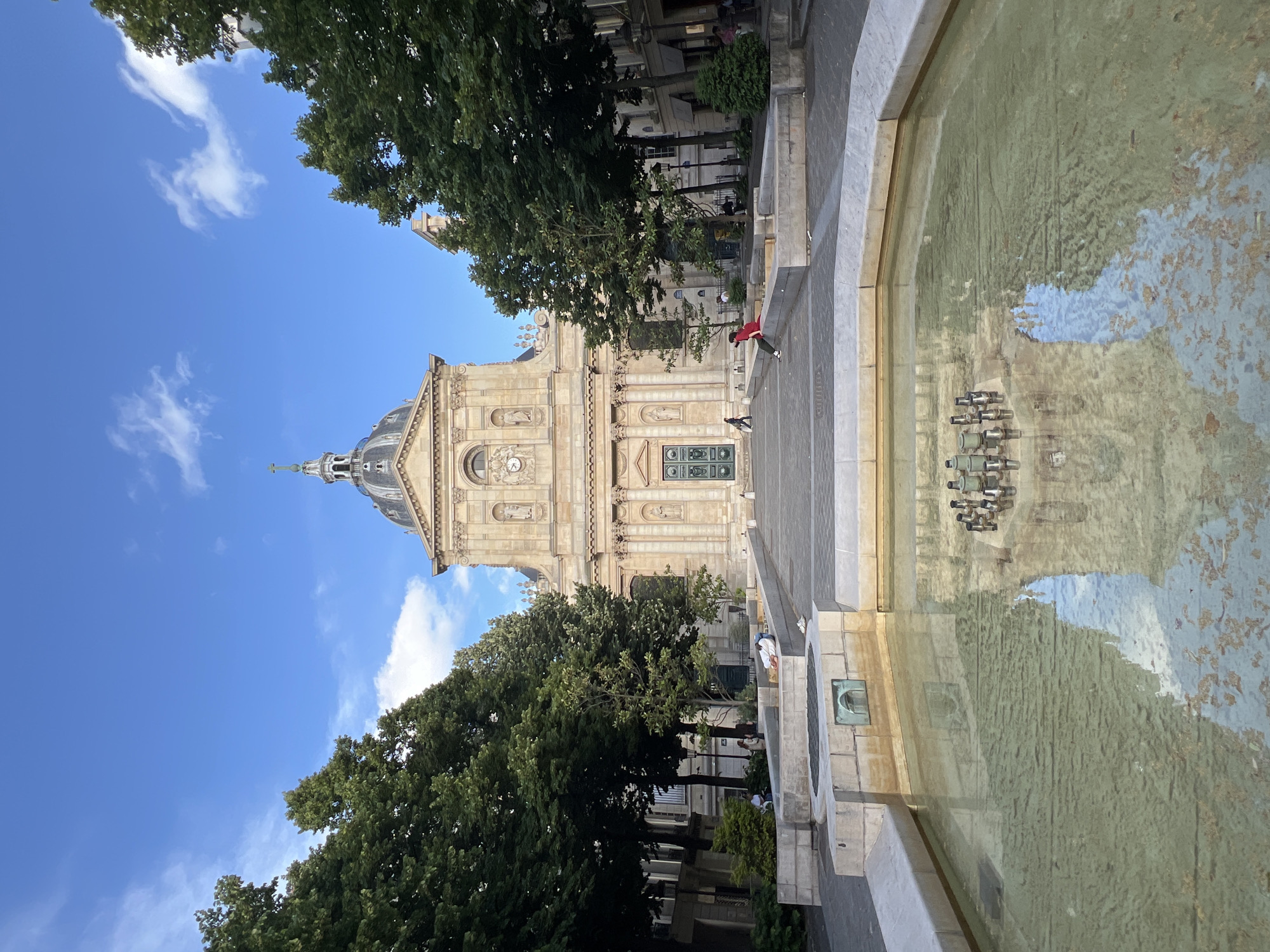 The image shows the exterior of the Saint-Étienne-du-Mont church in Paris, France. The church features a classical facade with columns and a dome, and is flanked by trees. In the foreground, a fountain with clear water reflects the church and the sky. Several people are visible in the scene. One person is sitting on the edge of the fountain, another is lying down on a bench nearby, and a few others are walking in the background. The sky is blue with scattered white clouds, and the sun appears to be shining, casting shadows. The overall atmosphere is serene and picturesque, suggesting a peaceful day in a public space.