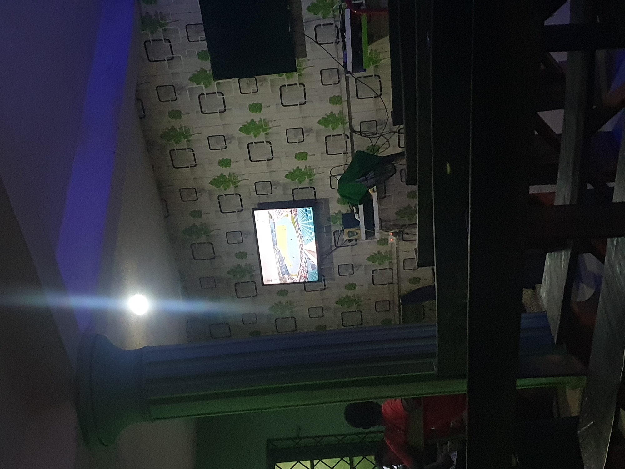 This image captures an indoor scene, likely in a casual lounge or common area, in Lokoja, Nigeria, under artificial lighting, suggesting nighttime or a low-light environment.
The central wall is covered with patterned wallpaper featuring light grey squares with dark outlines and scattered green, stylized leaf or tree motifs. A flat-screen television is mounted on this wall, displaying a brightly lit sports event, specifically what appears to be a football (soccer) match with a green field and stadium visible on screen. Above and to the right of the television, numerous black cables run along the wall, connecting to several electronic components, including a dark rectangular object mounted higher up and another piece of equipment covered by a green fabric.
On the far left, a bright light source, possibly a wall-mounted lamp or bulb, creates a strong glare. Below it, a segment of a light-colored, fluted column is visible. A strip of blue ambient lighting illuminates the upper left portion of the wall.
To the right, a dark-colored staircase, possibly made of wood or metal, dominates a significant portion of the frame, showing its treads and risers. In the bottom-right corner, a person wearing a red shirt is partially visible, bent over near a window or opening with a dark, grilled pattern.
The overall atmosphere suggests a relaxed setting, possibly for watching sports. There is no visible text in the image.