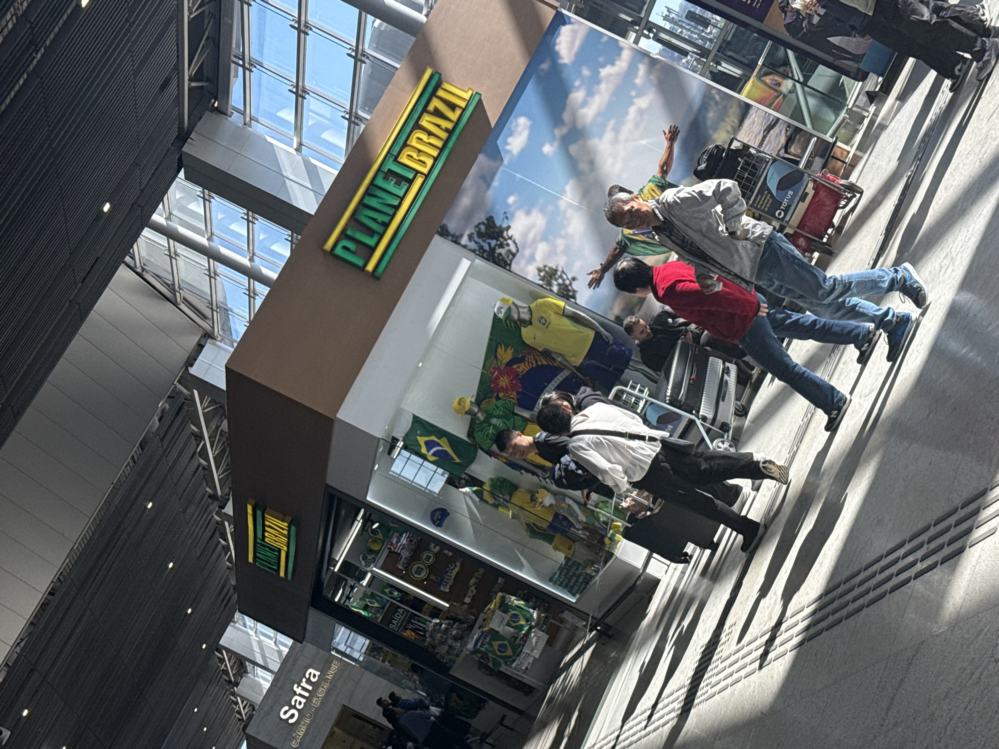 The image depicts a bustling indoor scene within what appears to be a modern airport terminal, specifically identified as Guarulhos International Airport (GRU) in Guarulhos, Brazil. The primary visual elements include two retail establishments: "PLANET BRAZIL," a souvenir shop prominently featuring Brazilian national colors and merchandise, and "Safra CÂMBIO - EXCHANGE," a currency exchange service.
The "PLANET BRAZIL" shop displays numerous Brazilian flags, yellow and green sports jerseys on mannequins, hats, and other themed souvenirs within its glass storefront. Above the shop, and extending onto a large wall graphic, is its distinctive green and yellow signage. The adjacent "Safra CÂMBIO - EXCHANGE" sign is visible to the left.
In the foreground and midground, several individuals are in various stages of transit or browsing. A man wearing a light grey jacket, blue jeans, and dark athletic shoes stands centrally, looking towards the camera with a slight smile. To his immediate left, another man in a red jacket and blue jeans is partially visible, facing away. Further to the left, a person in a white long-sleeved shirt and dark pants is seen from behind, pulling a black wheeled suitcase. A red luggage cart with "TOTUS" branding is visible behind the central figures. The background includes a large graphic of a person with outstretched arms against a cloudy sky, beneath an upper "PLANET BRAZIL" sign.
The environment features a high, architecturally complex ceiling with exposed metal framework and integrated lighting fixtures. Large, multi-paneled glass windows form portions of the walls and potentially the roof, allowing abundant natural sunlight to penetrate the interior. This strong illumination creates distinct, elongated shadows of people and objects on the light-colored, tiled floor. The floor itself incorporates tactile paving strips, consisting of raised rectangular blocks, indicating pathways. Multiple other individuals are visible throughout the scene, some browsing inside the "PLANET BRAZIL" shop or walking in the background.
